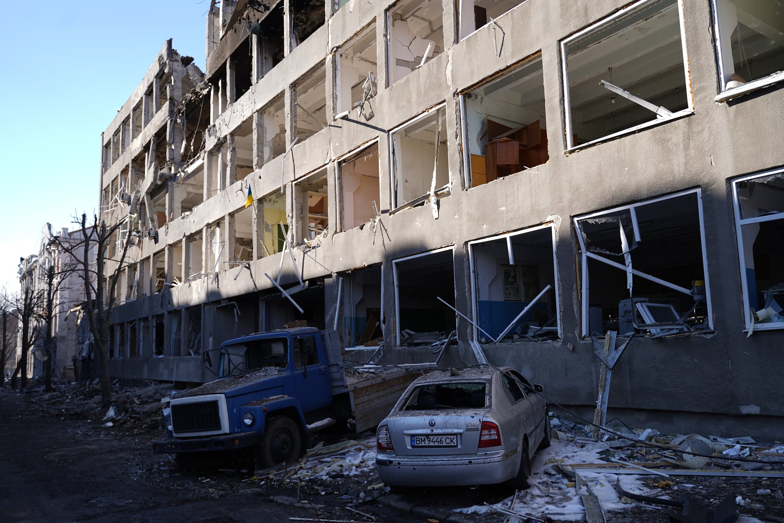 Разрушенное в результате боевых действий многоэтажное здание с выбитыми окнами. На переднем плане — синий грузовик и серебристый легковой автомобиль, поврежденные обломками. Из одного из окон здания виден маленький украинский флаг.