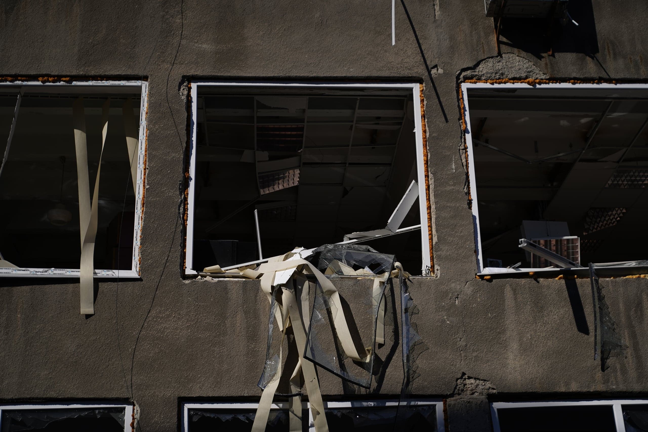 Крупный план темного фасада здания с тремя разбитыми окнами. Из центрального оконного проема свисают осколки стекла и длинные бежевые полосы, внутри виден разрушенный подвесной потолок.