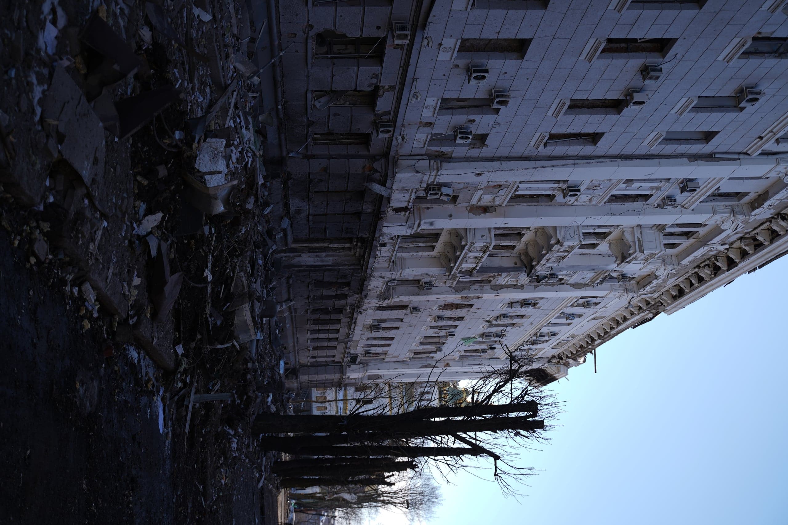 Разрушенный обстрелами многоэтажный дом с изрешеченным фасадом и выбитыми окнами. Перед зданием лежат груды темных строительных обломков и мусора. Справа виден ряд обрезанных голых деревьев на фоне ясного голубого неба.