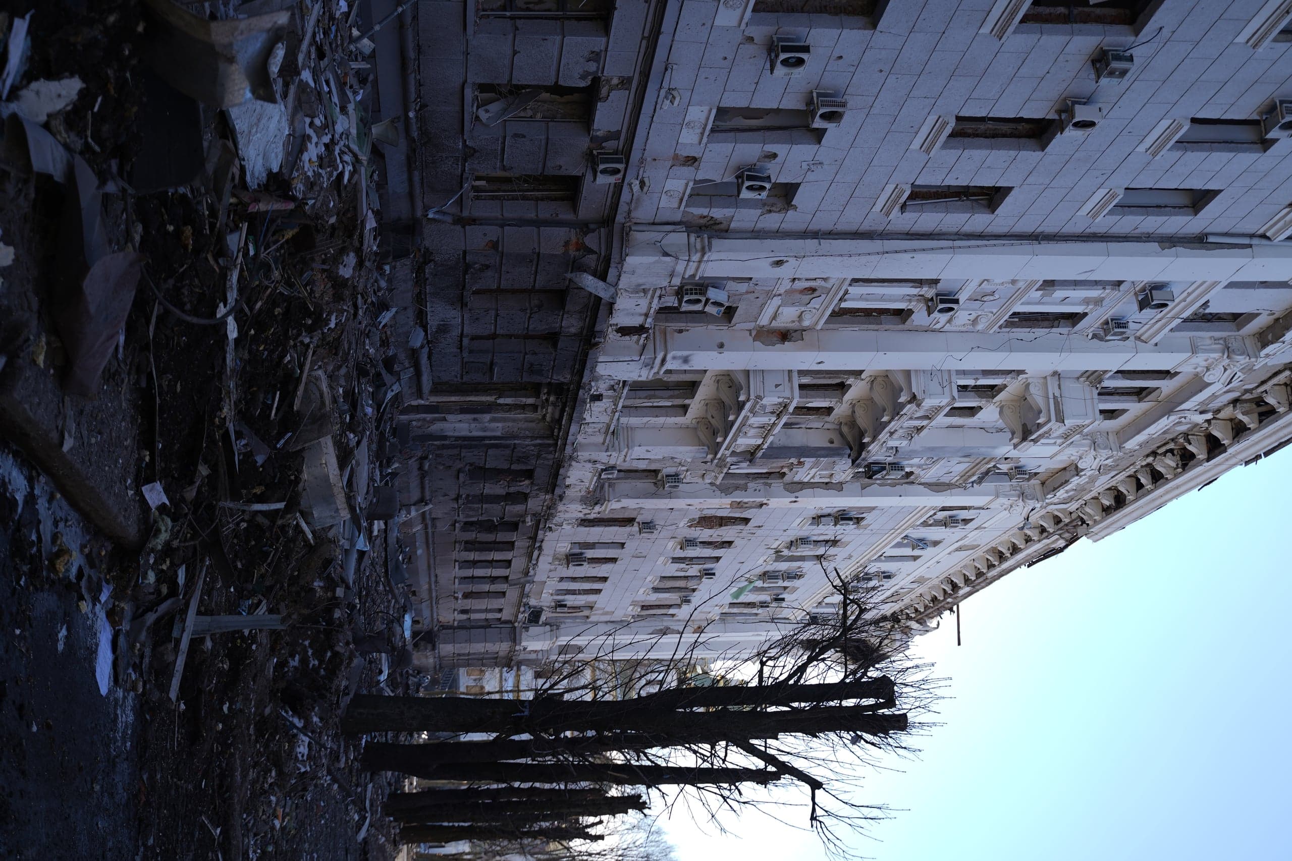 Фасад сильно поврежденного обстрелами многоэтажного здания с разбитыми окнами и разрушенными балконами. Перед домом — большая груда строительного мусора и ряд обрезанных деревьев.