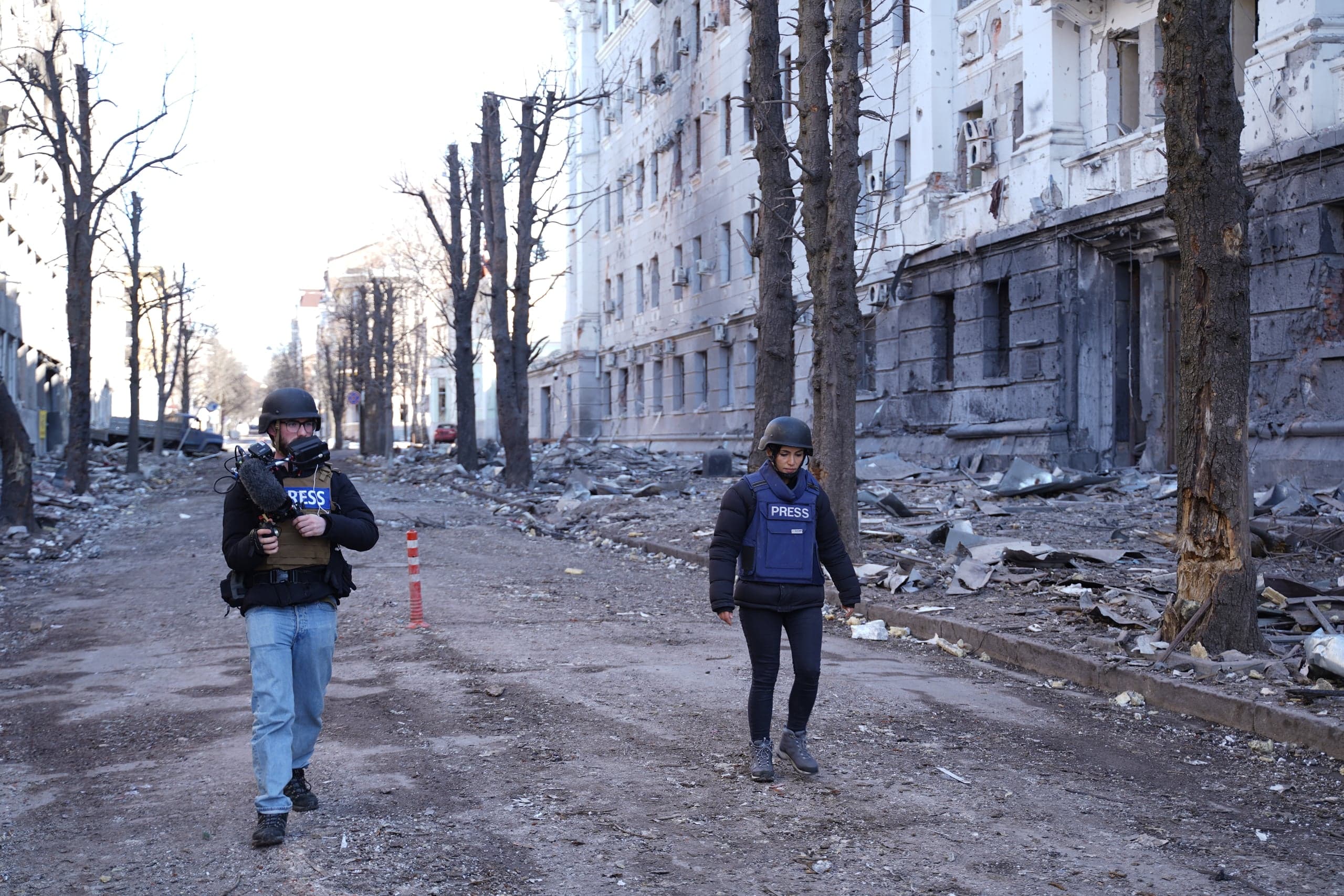 Двое военных корреспондентов в жилетах "PRESS" и касках идут по разрушенной, засыпанной обломками городской улице на фоне сильно поврежденного здания.
