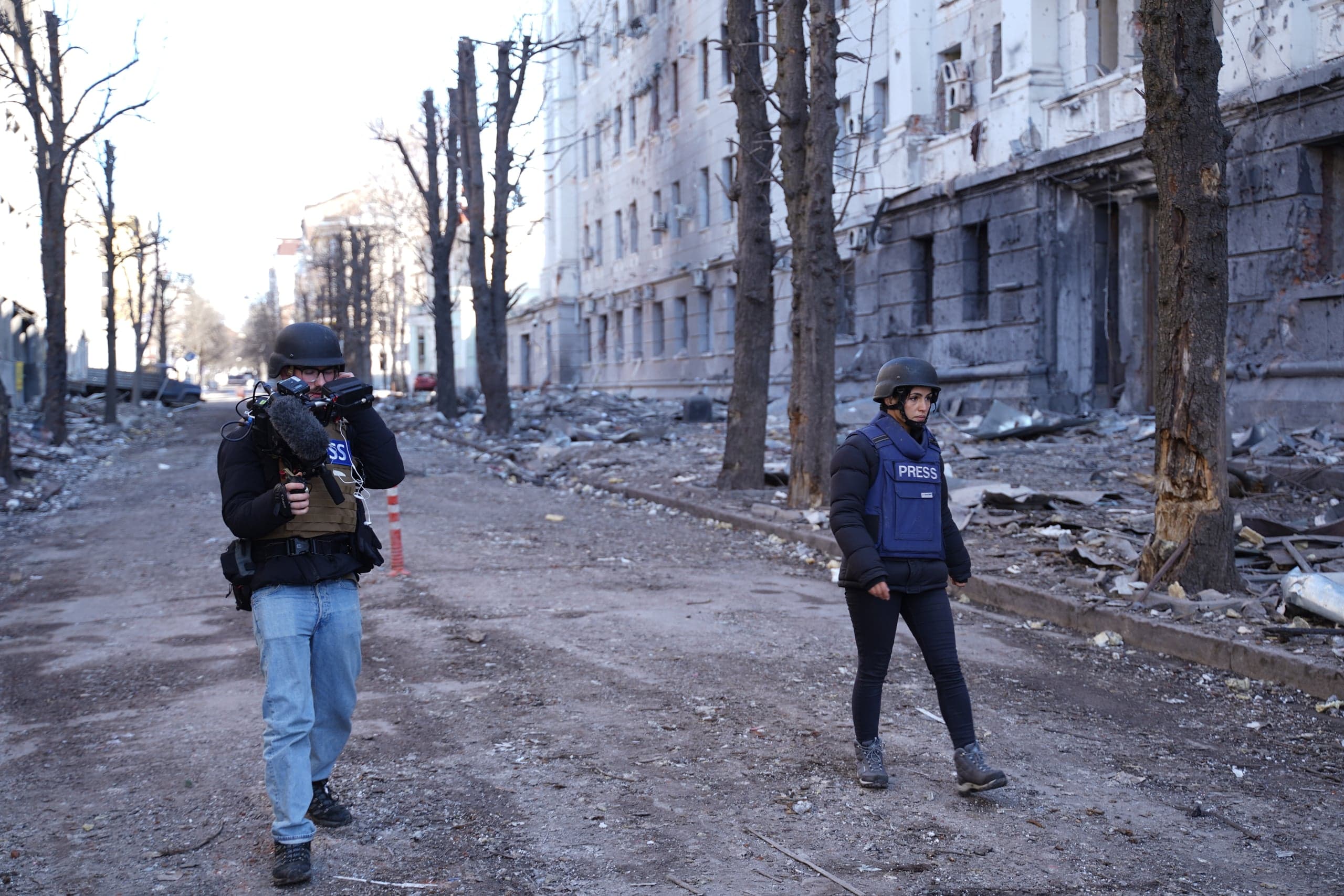 Двое журналистов в касках и бронежилетах (женщина в синем жилете "PRESS") идут по разрушенной обломками улице на фоне сильно поврежденного здания.