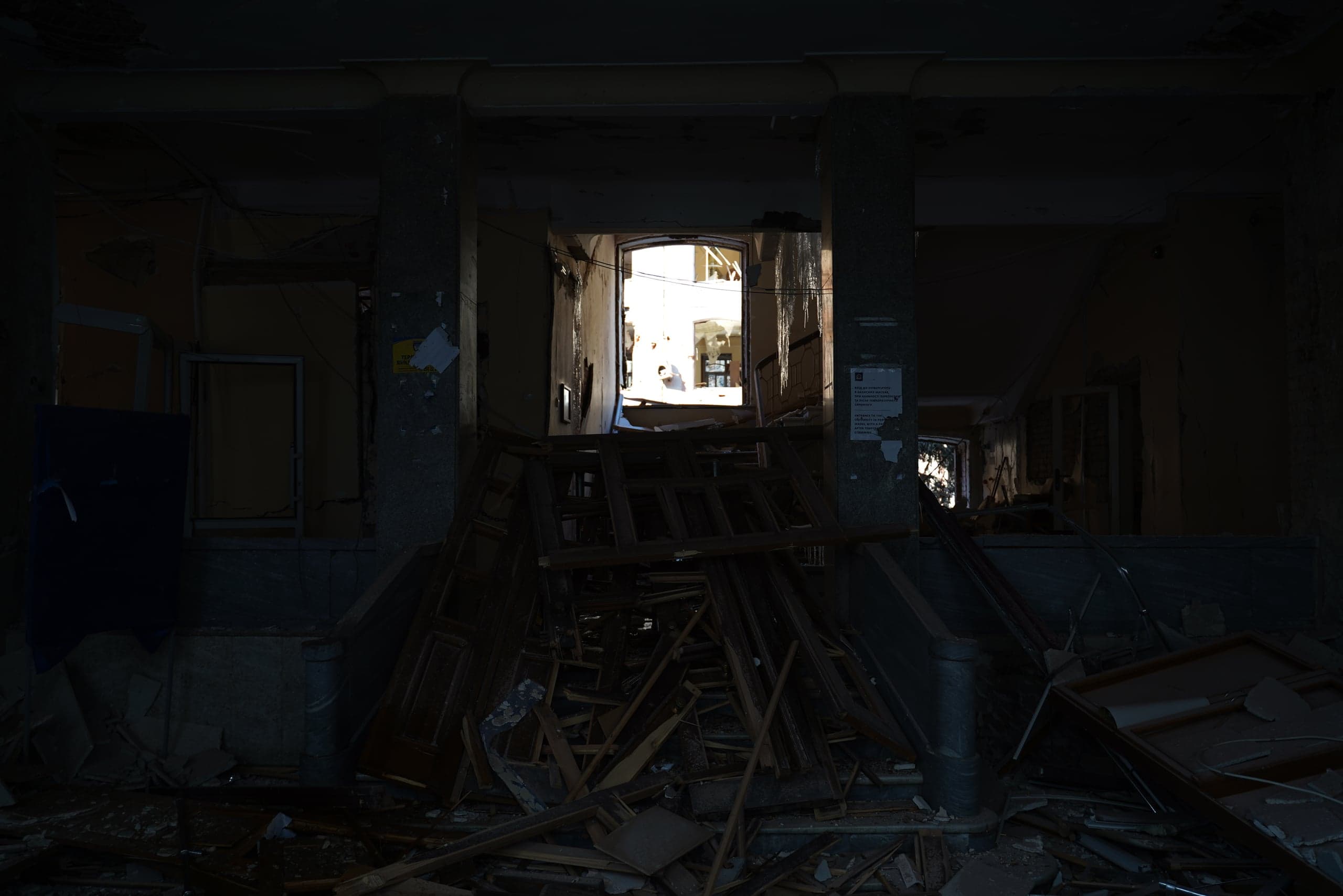Темный интерьер разрушенного здания. Груды деревянных рам и обломков перед двумя колоннами. Яркий дневной свет пробивается через центральный проем на заднем плане.