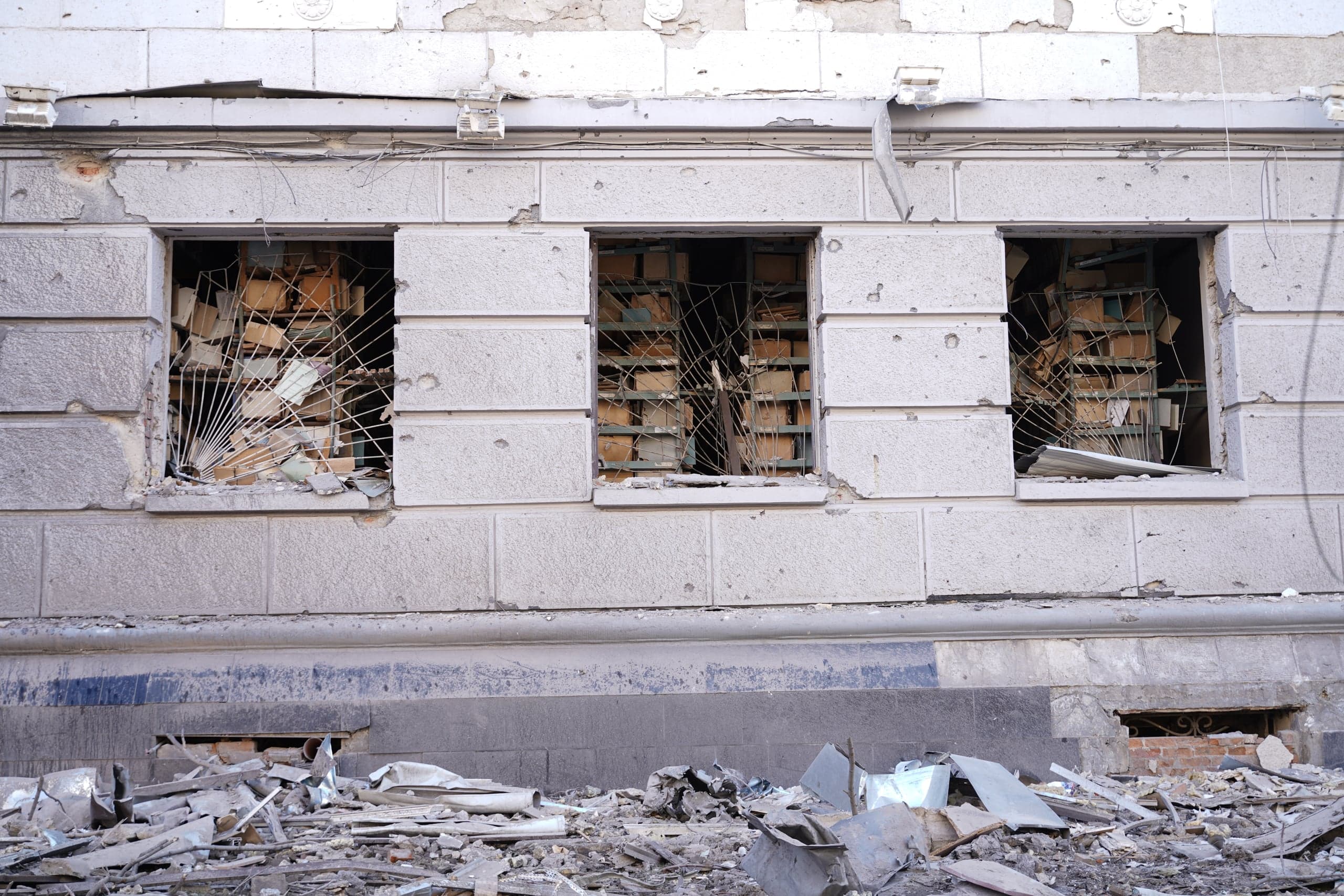 Фасад разрушенного здания с тремя выбитыми окнами, за которыми видны стеллажи, заваленные документами и коробками. Перед стеной лежит большая груда строительного мусора.