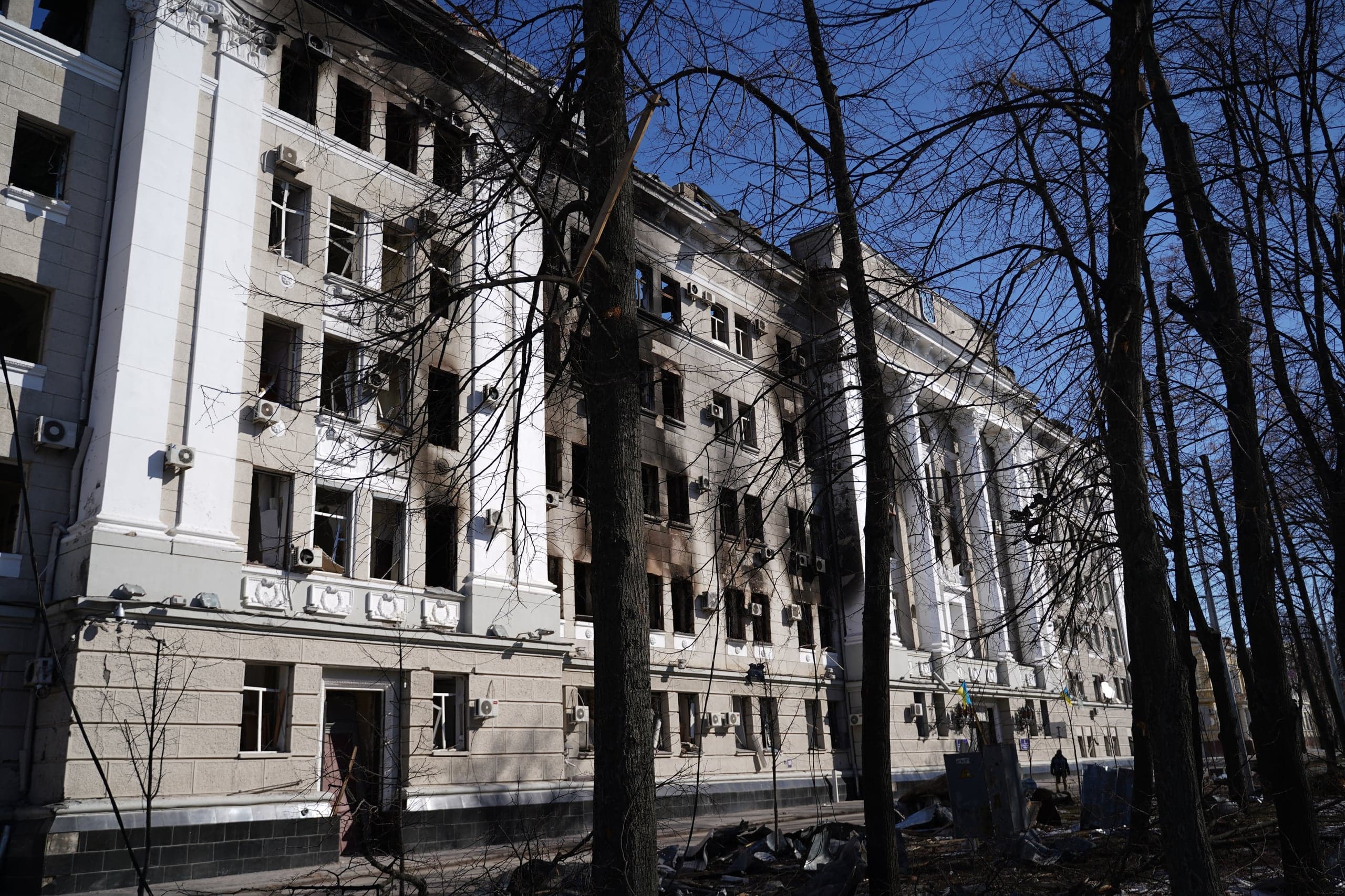 Поврежденное, закопченное огнем многоэтажное здание классического стиля с выбитыми окнами, снятое сквозь силуэты голых деревьев на фоне синего неба.