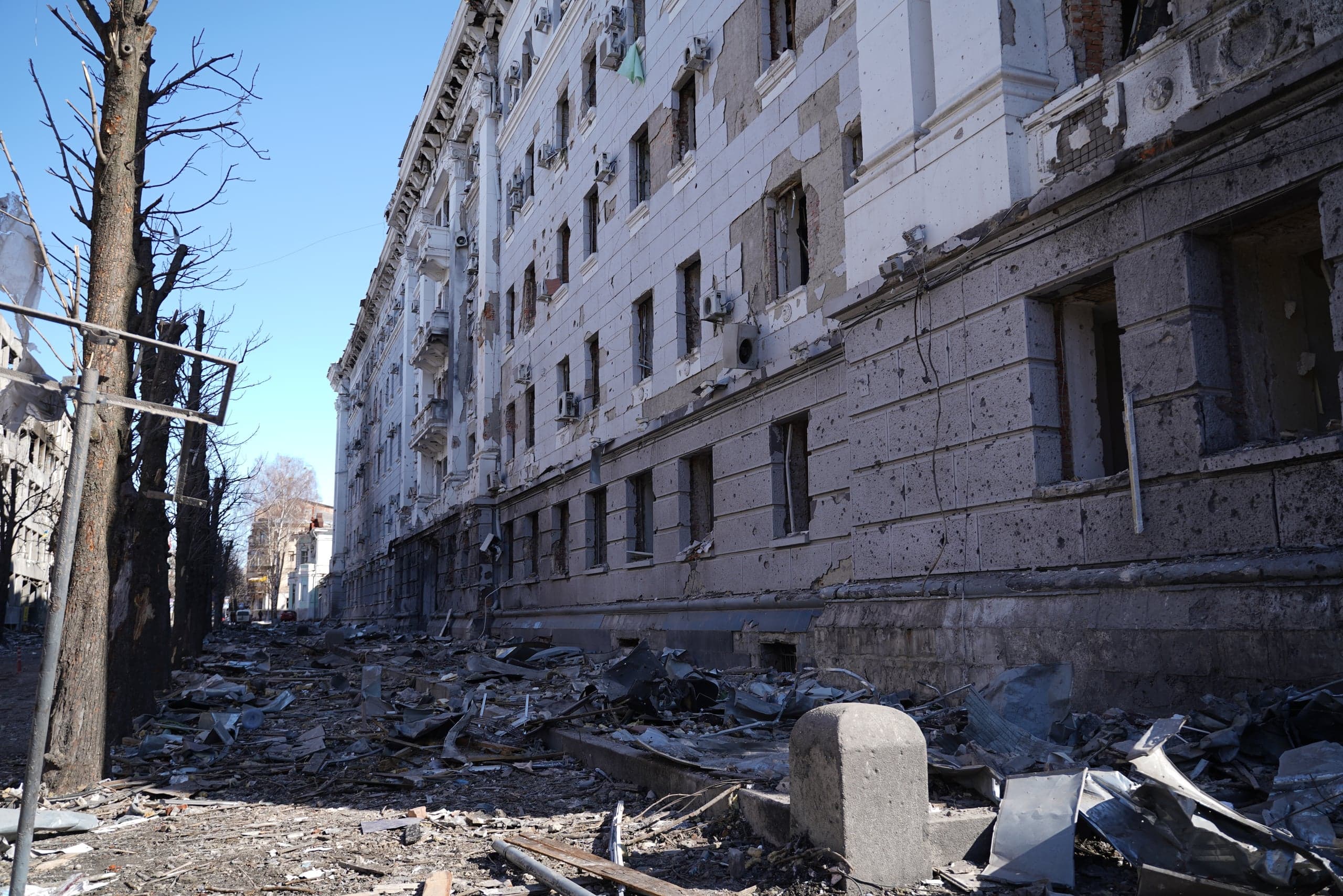 Сильно поврежденное многоэтажное здание с фасадом, испещренным отверстиями от снарядов. Перед зданием — обширные завалы мусора, обломков и искореженного металла. Слева видны голые стволы деревьев.