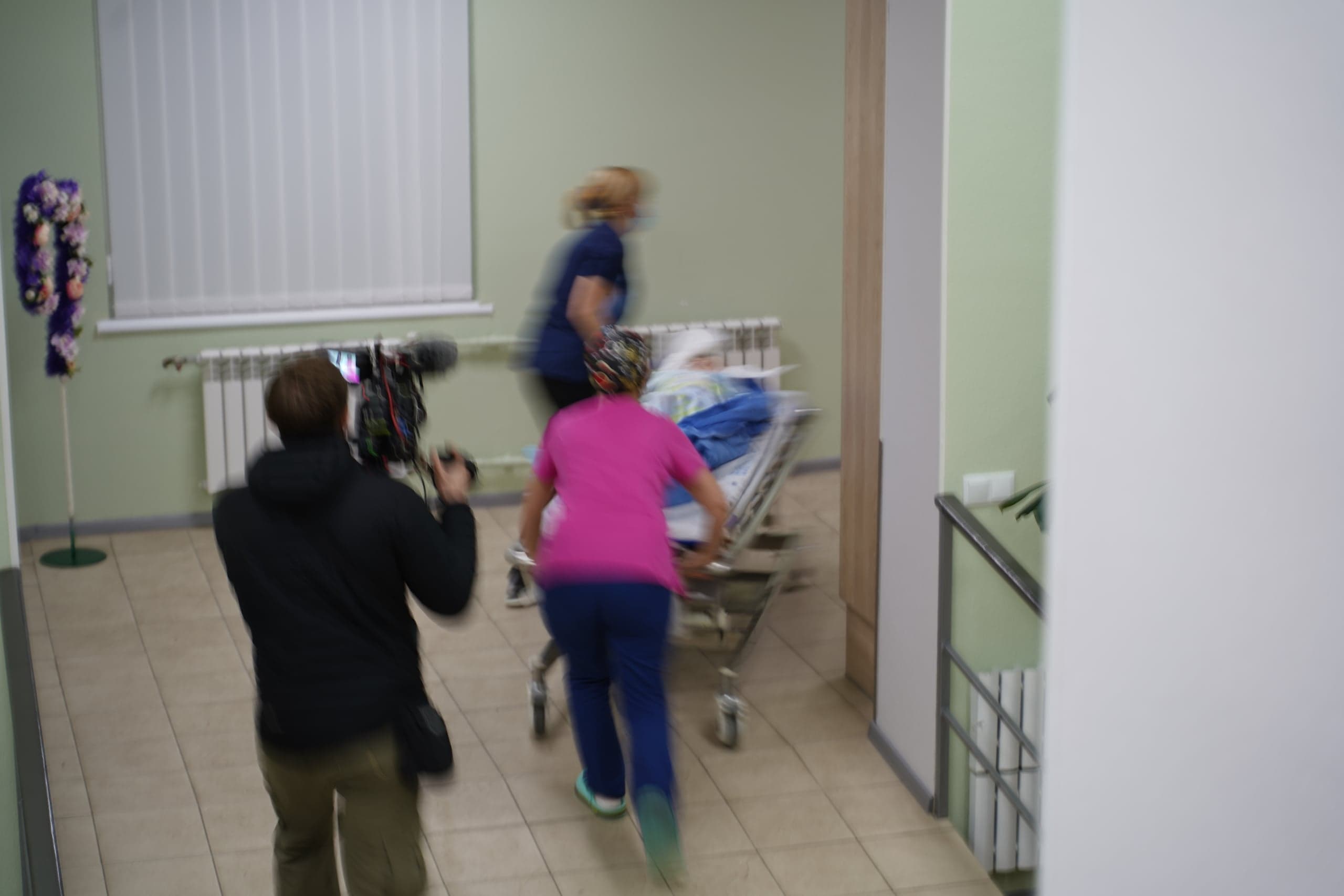 Оператор снимает двух медработников, быстро везущих пациента на каталке в больничном коридоре. Снимок размыт из-за движения.