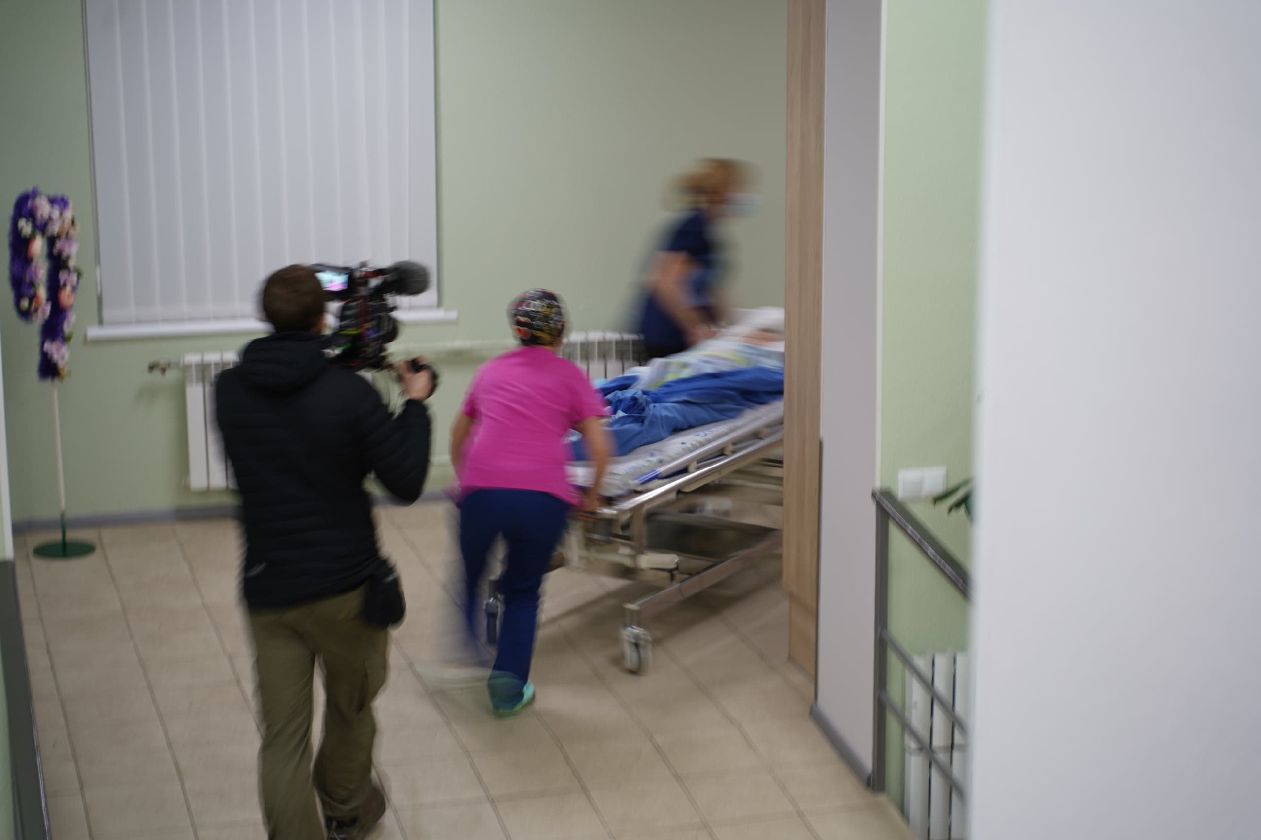 Оператор с камерой снимает, как медперсонал торопливо везет пациента на каталке по больничному коридору (кадр с сильным смазыванием движения).