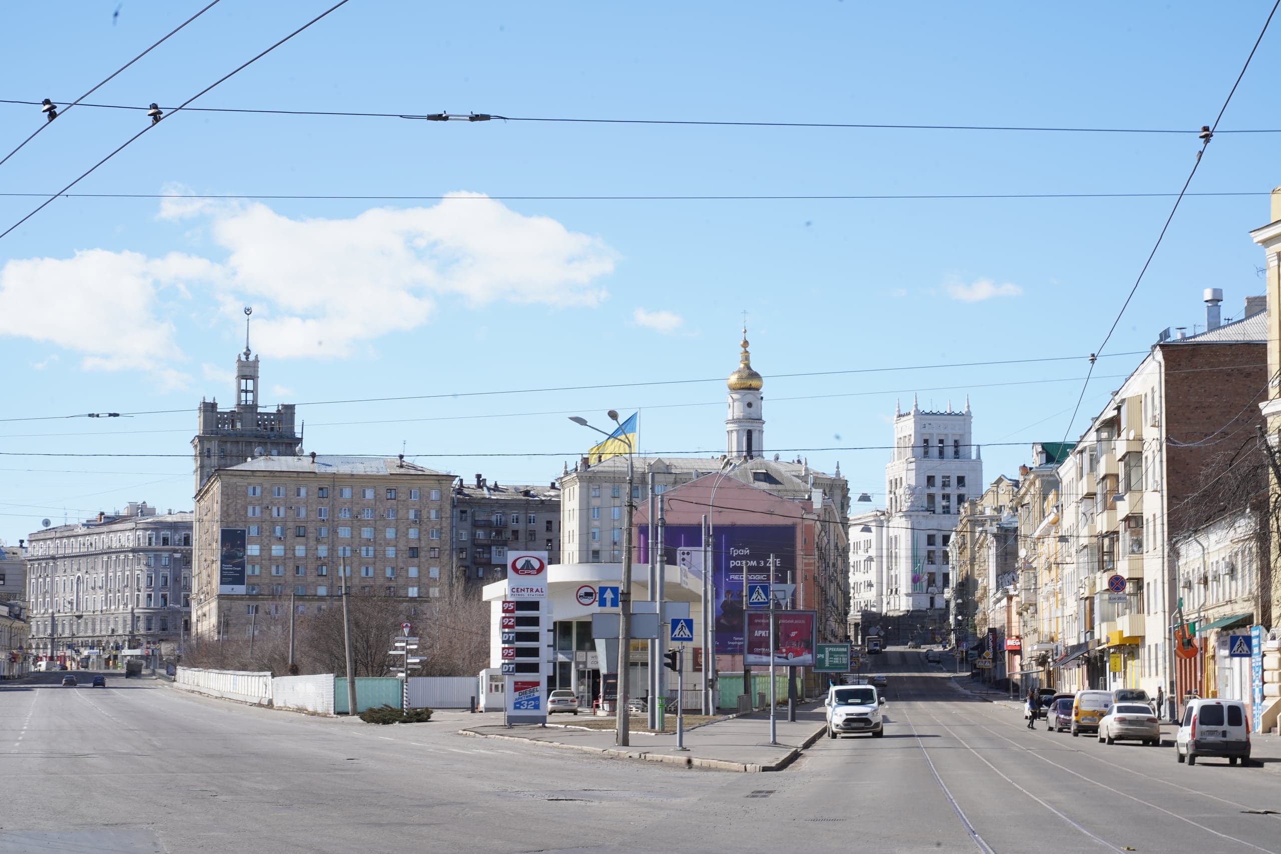 Панорама широкой городской улицы в солнечный день. На заднем плане доминируют исторические здания: серая сталинская башня, белая колокольня с золотым куполом и современная АЗС на углу. Дорога пересечена проводами для электротранспорта.