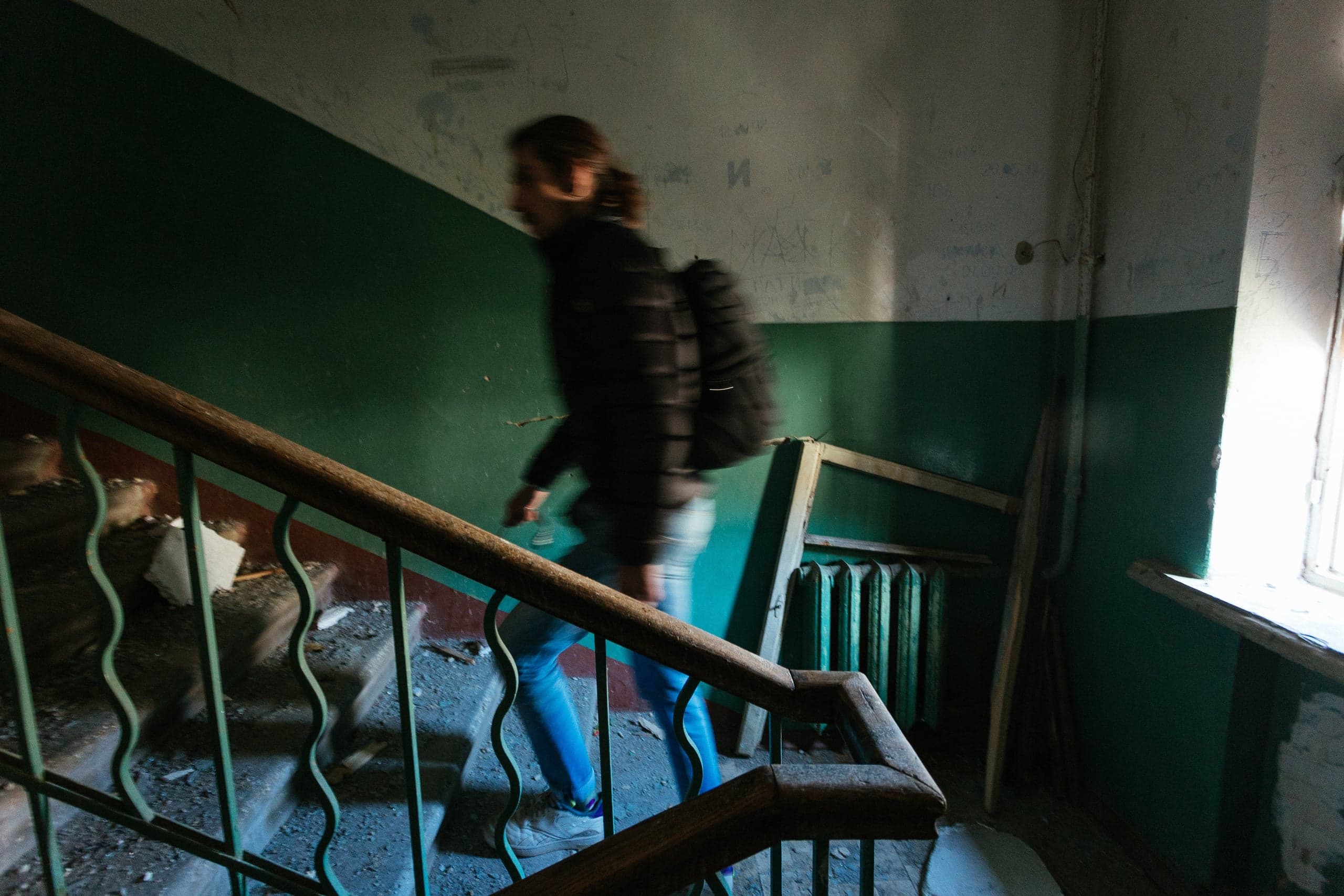 Размытый снимок человека с рюкзаком, поднимающегося по обветшалой лестнице с деревянными перилами в заброшенном помещении. Стены окрашены в темно-зеленый и грязно-белый, справа яркий свет из окна.