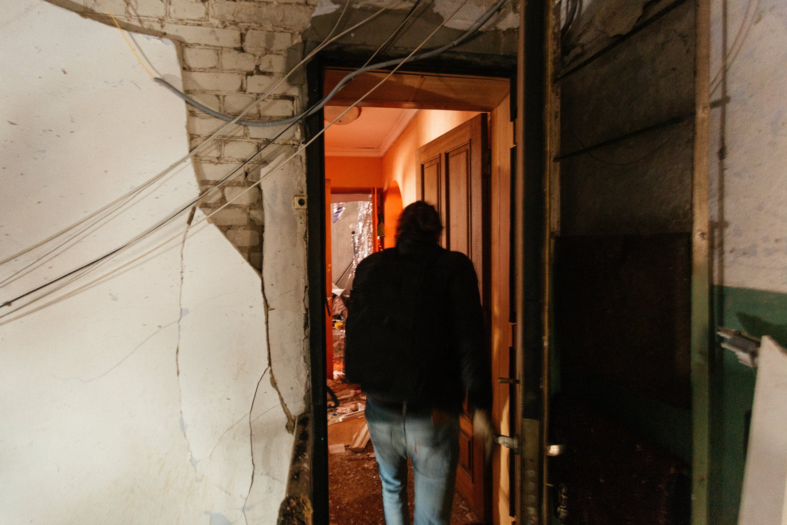 Человек со спины входит в дверной проем с разрушенными стенами и свисающими проводами. Интерьер освещен оранжевым светом, видны обломки.