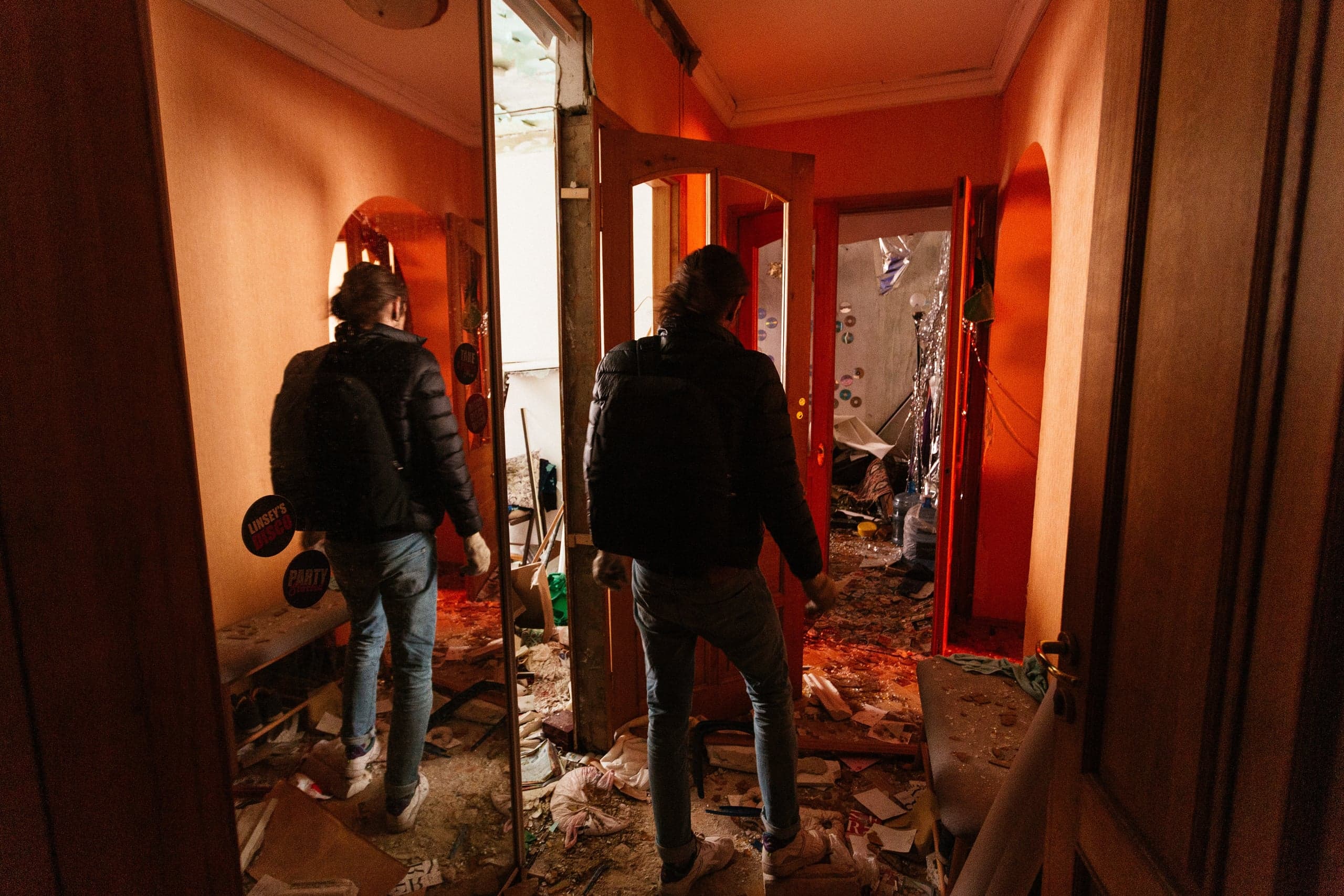 Мужчина в черной куртке и джинсах стоит посреди коридора разрушенного помещения с оранжевыми стенами. Пол усыпан мусором и обломками. В зеркале слева отражается его фигура и наклейки "LINDSEY'S" и "PARTY".