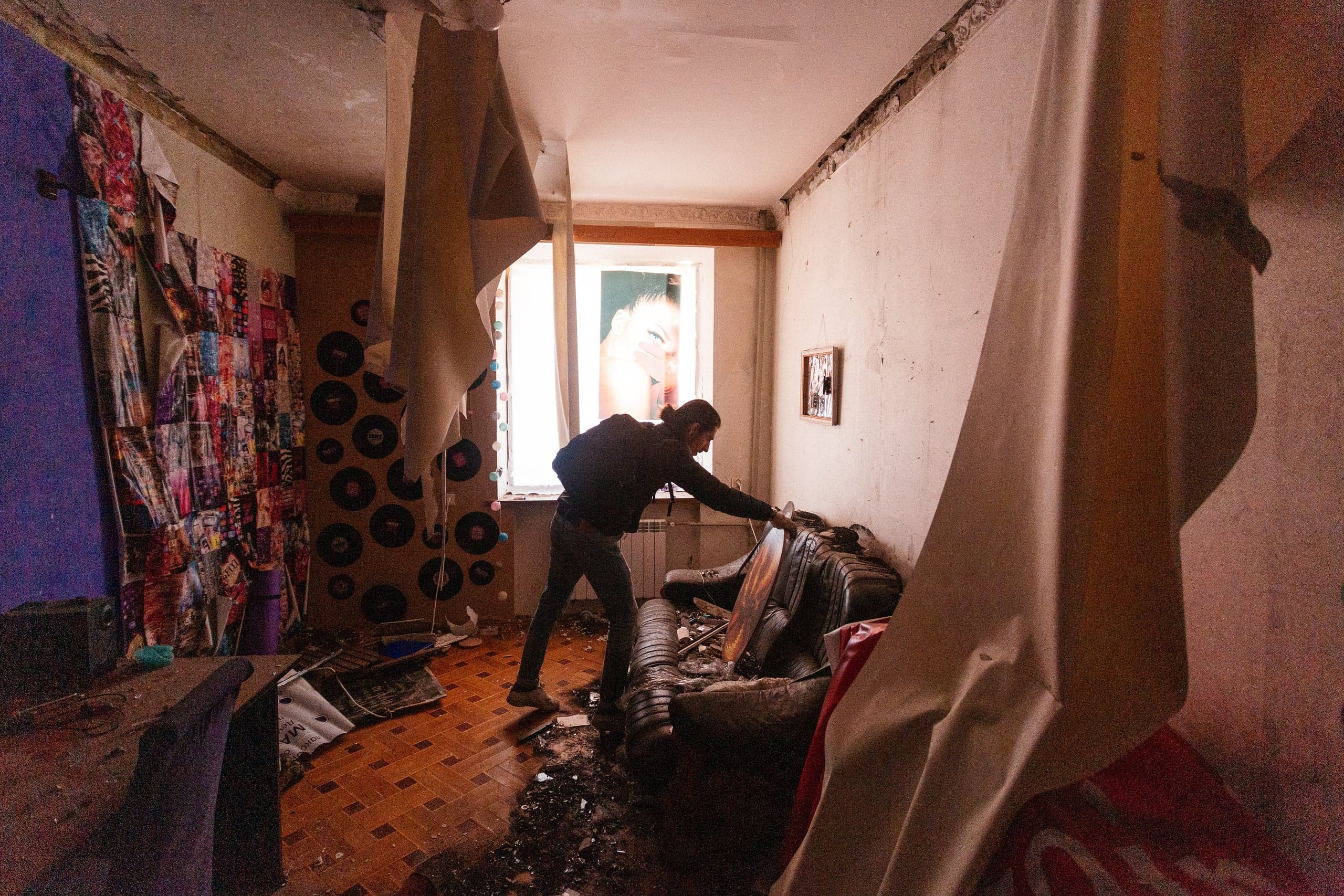 Мужчина осматривает разрушенный диван в сильно поврежденной комнате. Стены ободраны, пол покрыт сажей. В окне виден фрагмент постера.