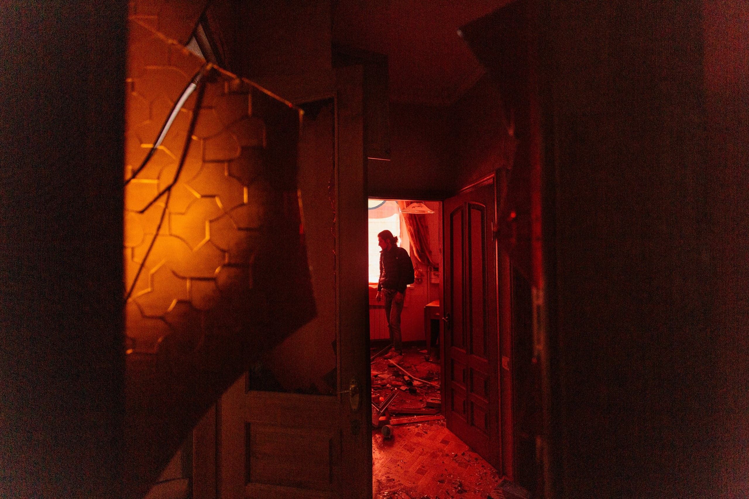 Снимок через дверной проем на разрушенное помещение в огненно-красных тонах, где человек стоит среди обломков на полу.