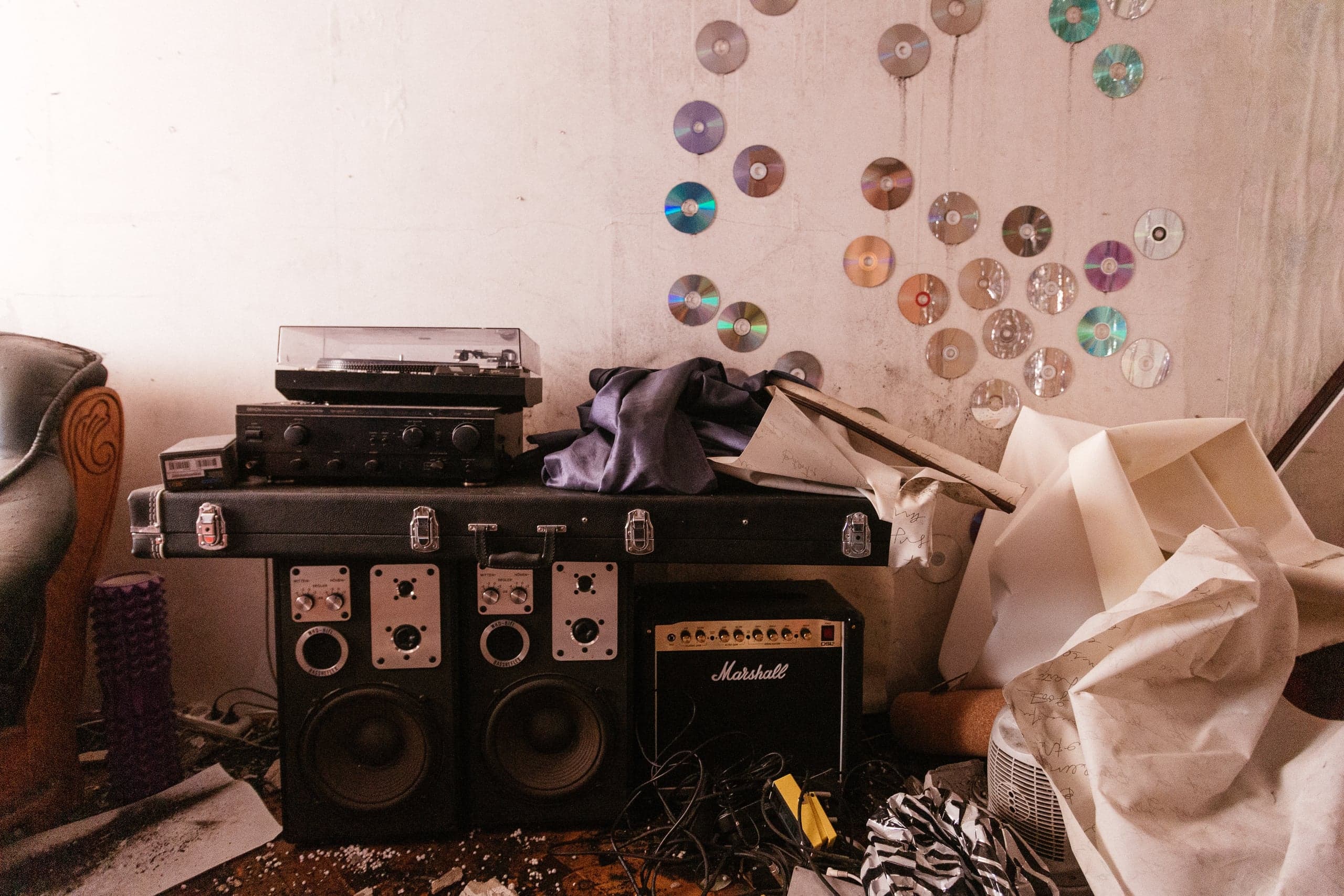Грязная комната с музыкальным оборудованием: проигрыватель и усилитель на кейсе, большие колонки и гитарный усилитель Marshall. К обшарпанной стене прикреплены многочисленные CD-диски в виде узора. Пол усеян проводами и скомканной бумагой.
