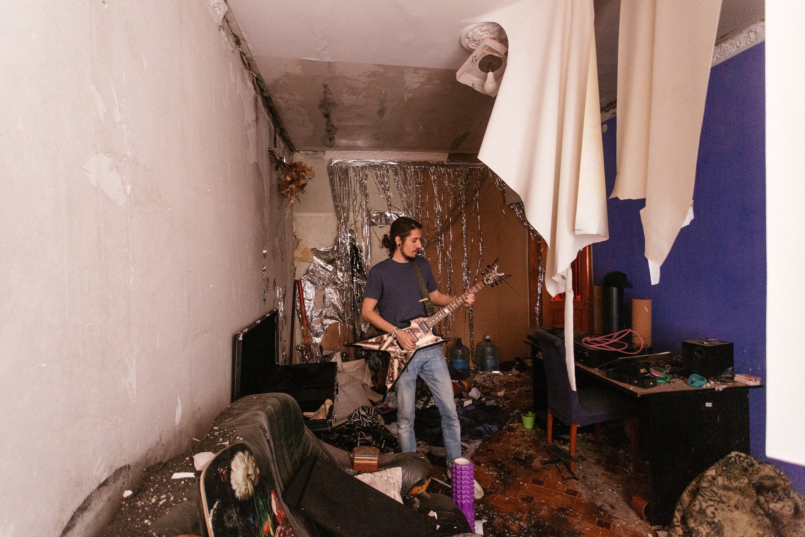 Музыкант с электрогитарой в хаотичной, разрушенной комнате, где обвалился потолок.