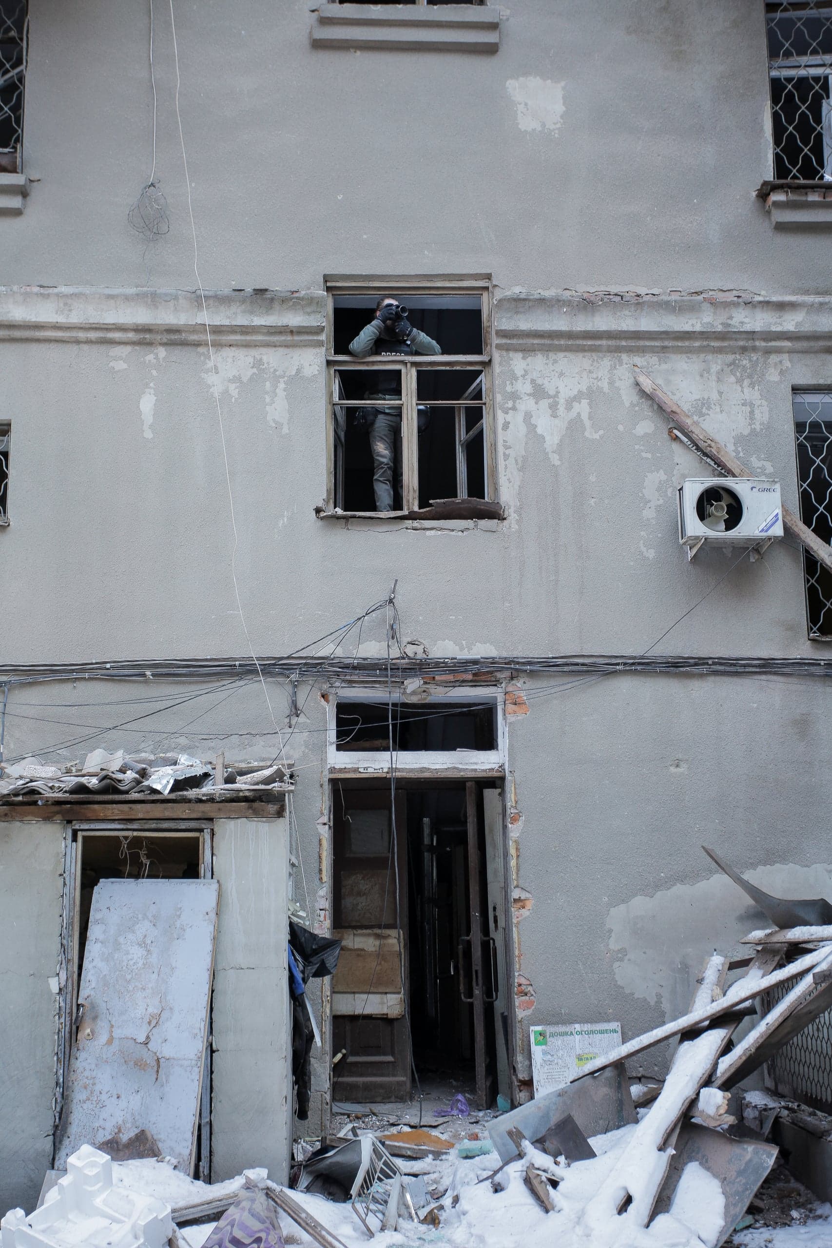 Поврежденный фасад здания; человек смотрит из разбитого окна, держа камеру; на земле завалы и снег.