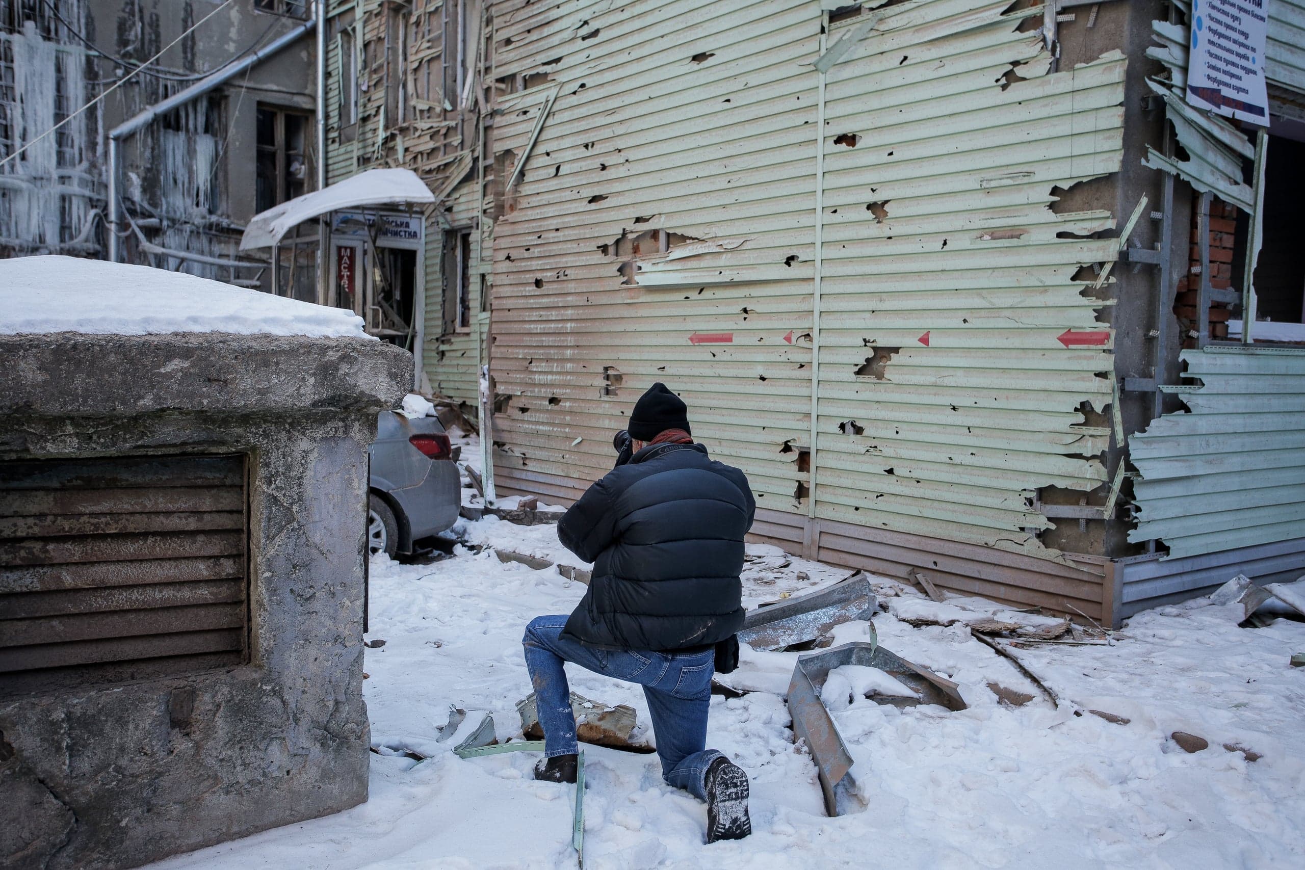 Фотограф стоит на коленях на снегу, снимая здание с многочисленными повреждениями от пуль и осколков.