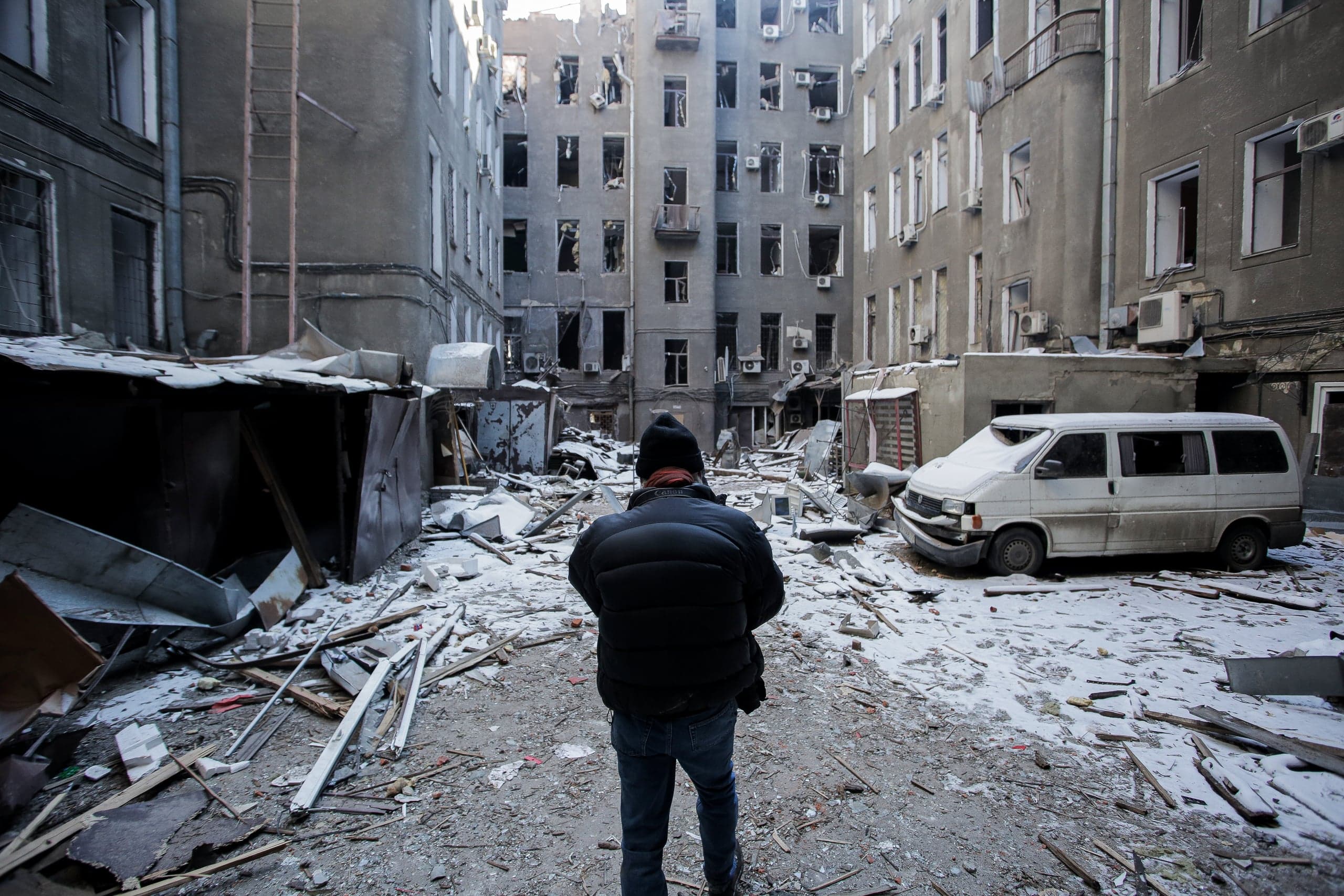 Мужчина стоит среди обломков и снега во дворе, окруженном сильно поврежденными обстрелом многоэтажными жилыми домами. Справа стоит разбитый фургон.