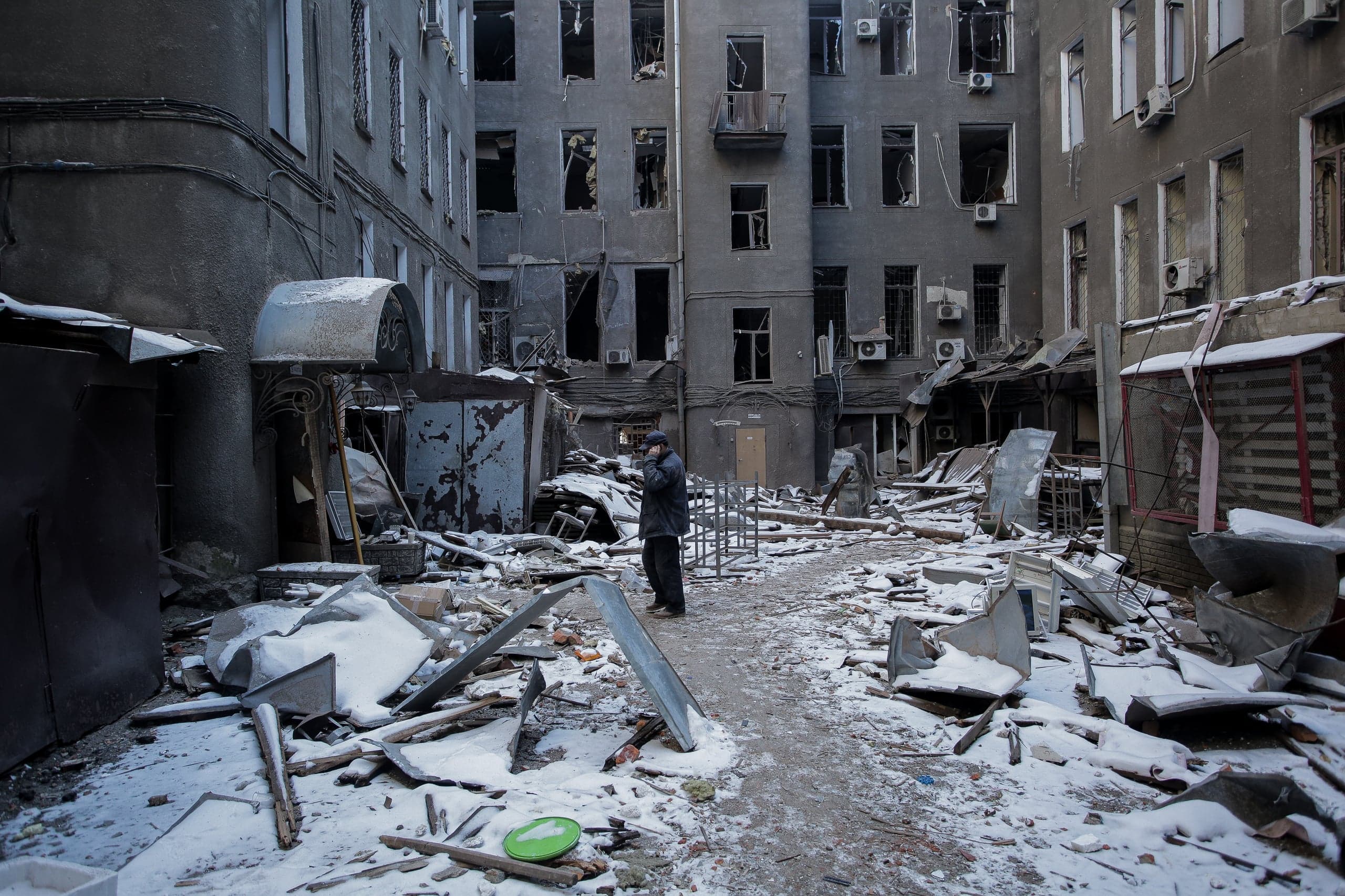 Внутренний двор разрушенного многоквартирного дома, усыпанный обломками и снегом. Пожилой человек стоит в центре двора. Окна здания выбиты.