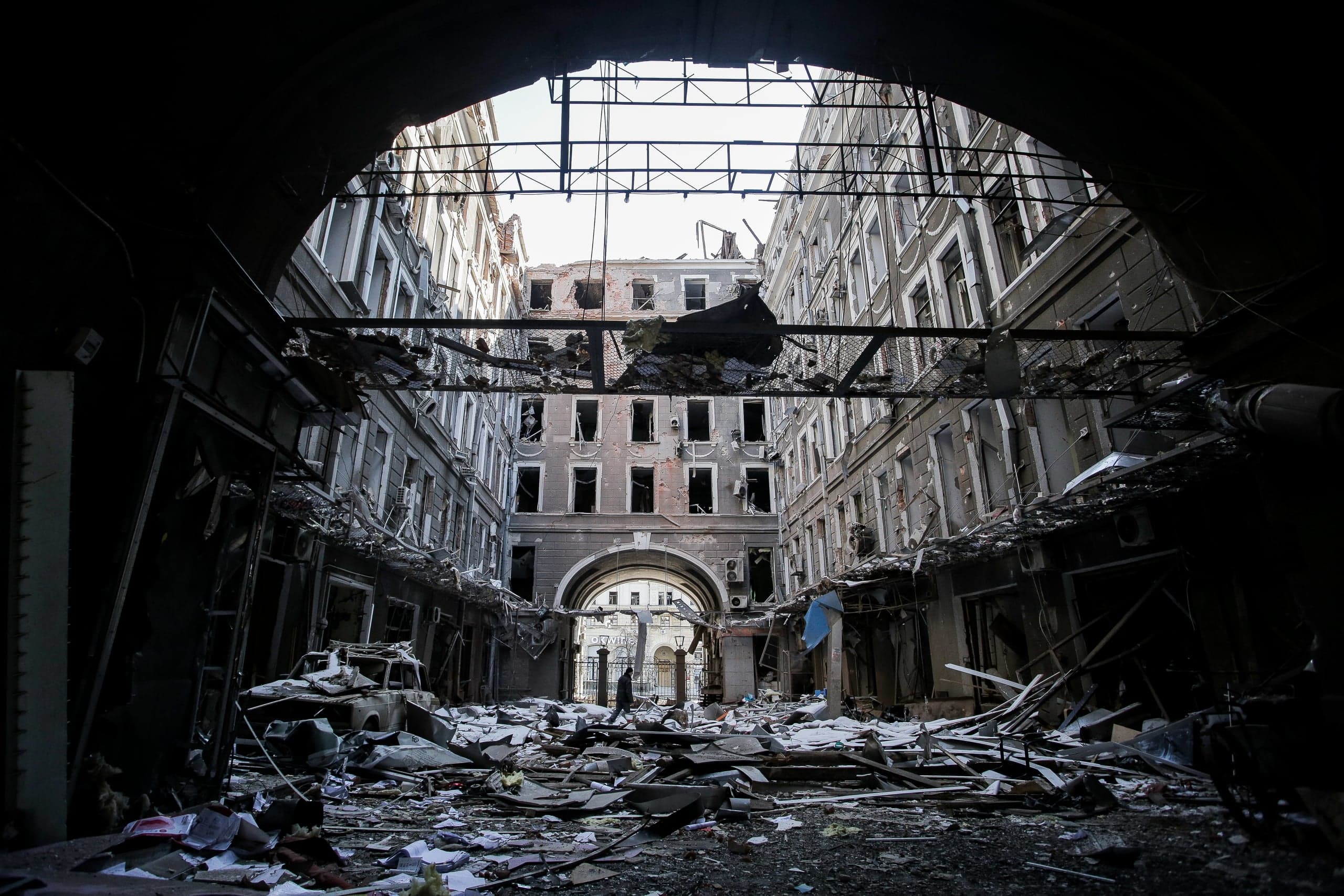 Разрушенный взрывом крытый проход (двор-колодец). Фасады зданий сильно повреждены, крыша обрушена, земля покрыта строительными обломками и мусором.