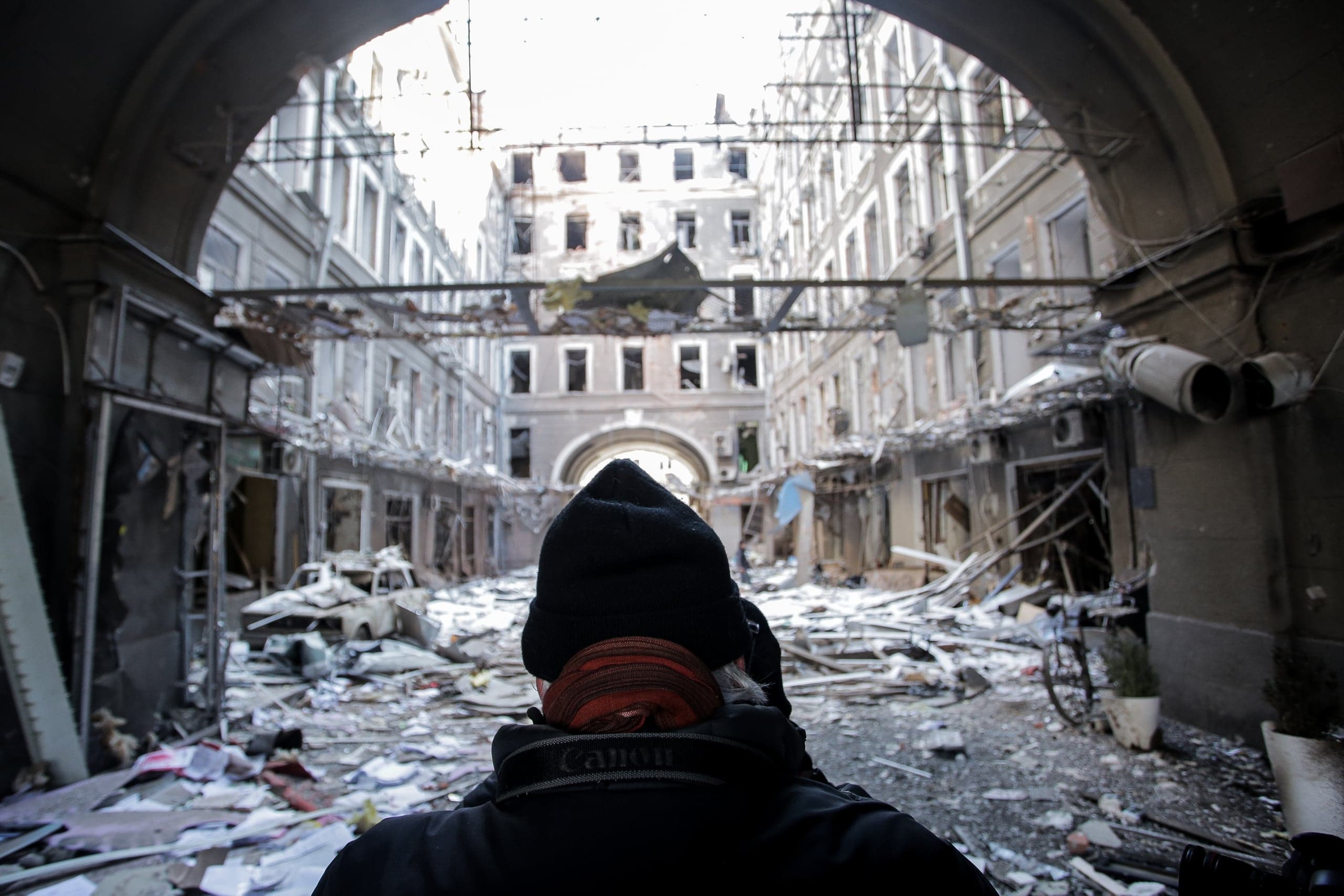 Человек смотрит через арку на разрушенный городской двор, заваленный обломками. Здания сильно повреждены, видны выбитые окна и разбитый автомобиль.