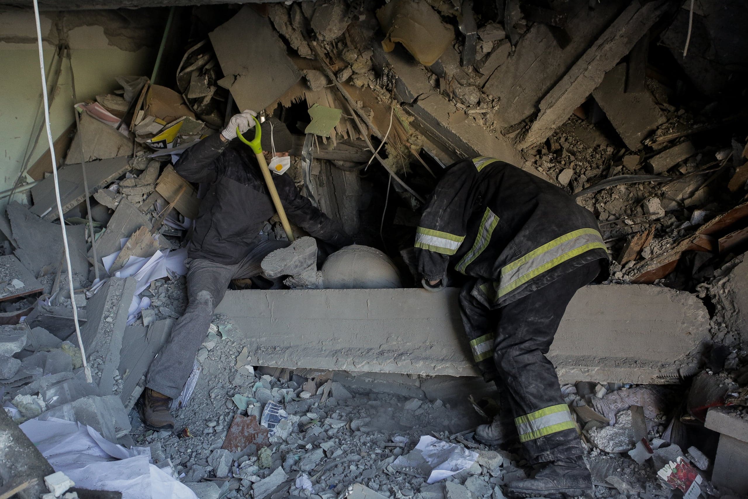 Спасатель и гражданский разбирают завалы разрушенного здания, используя лопату.