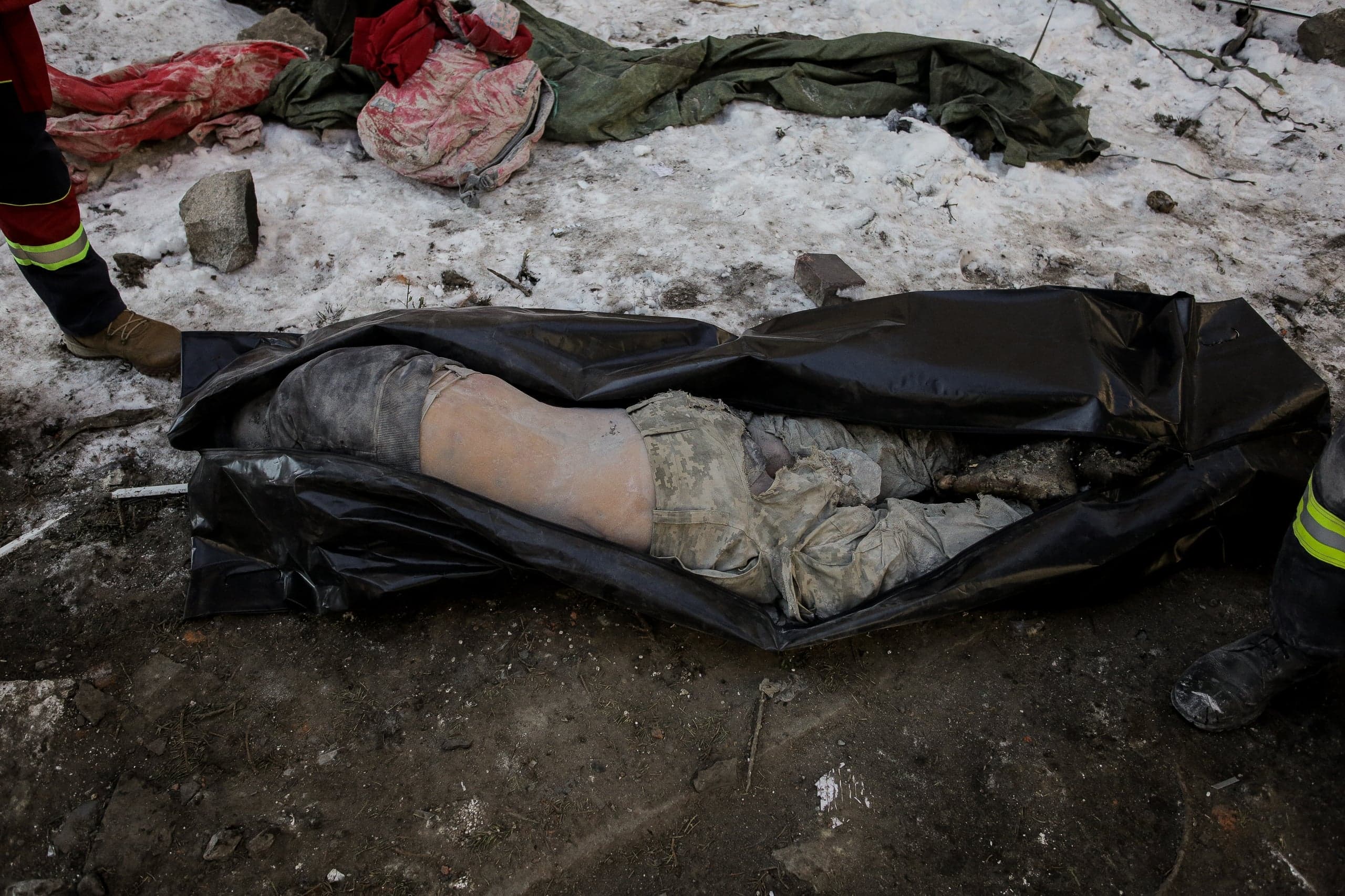 Труп человека в незакрытом черном пакете лежит на асфальте, рядом видно несколько других людей, на фоне снег