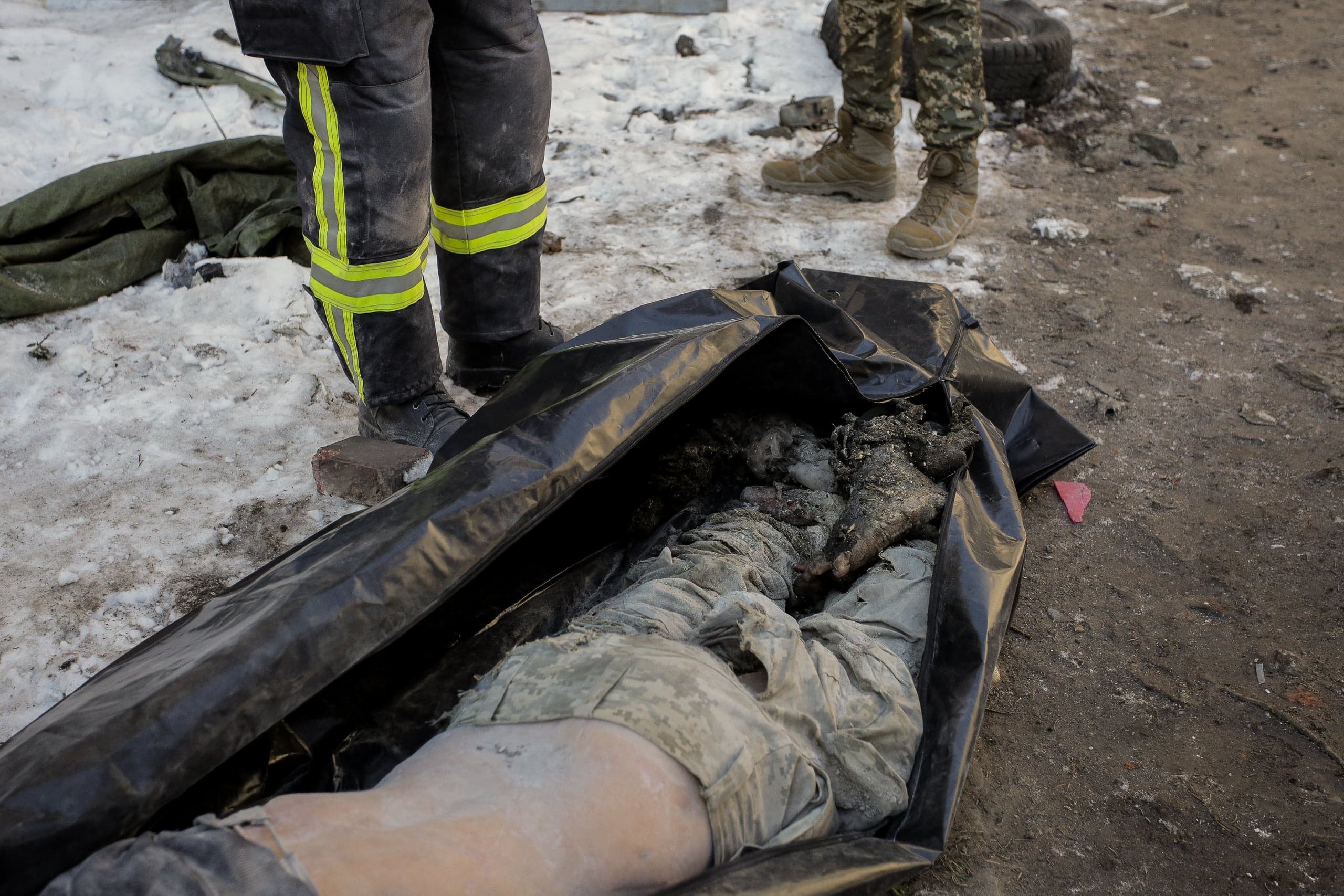Частично накрытое черным мешком тело (в камуфляже) на земле. Рядом видны ноги спасателя и военного.