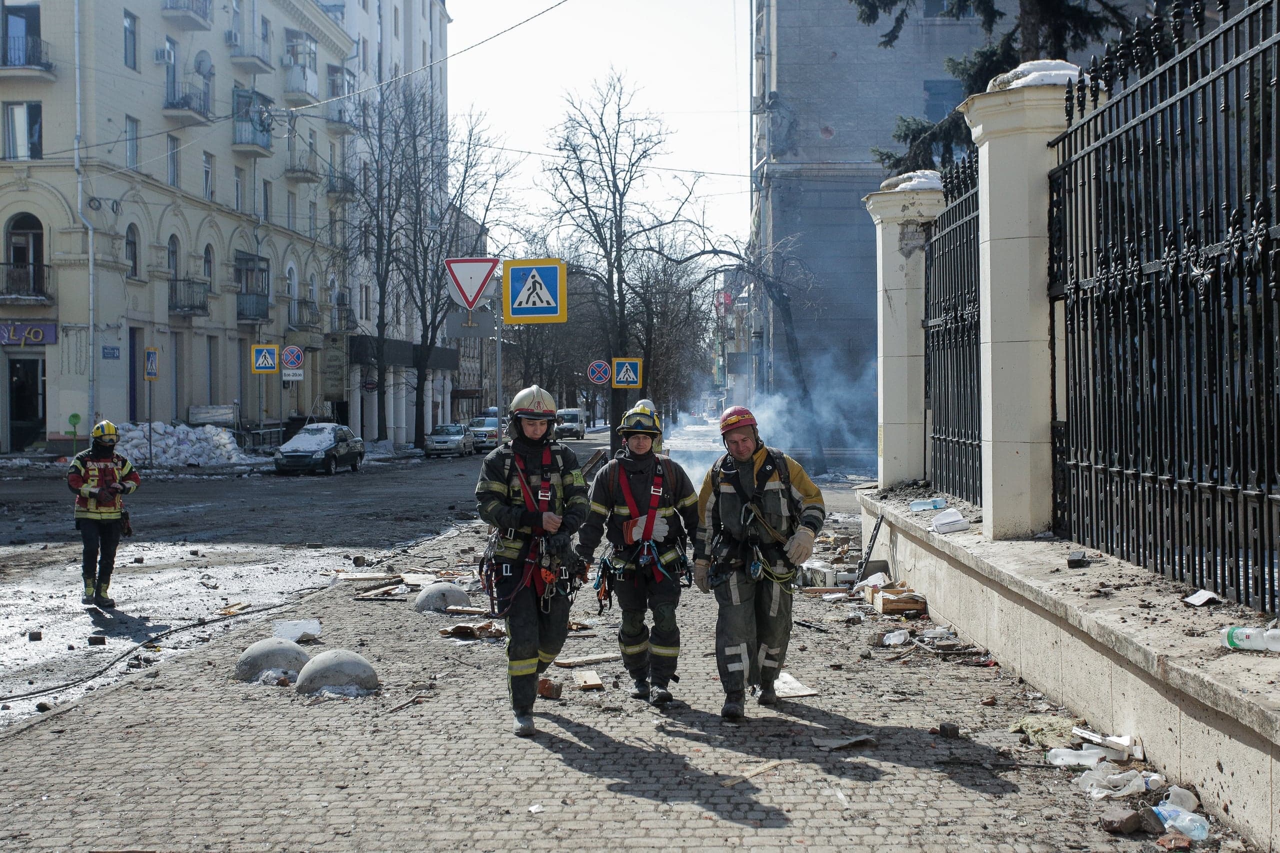 Три спасателя идут по разрушенной городской улице, усыпанной обломками. На заднем плане виден дым.