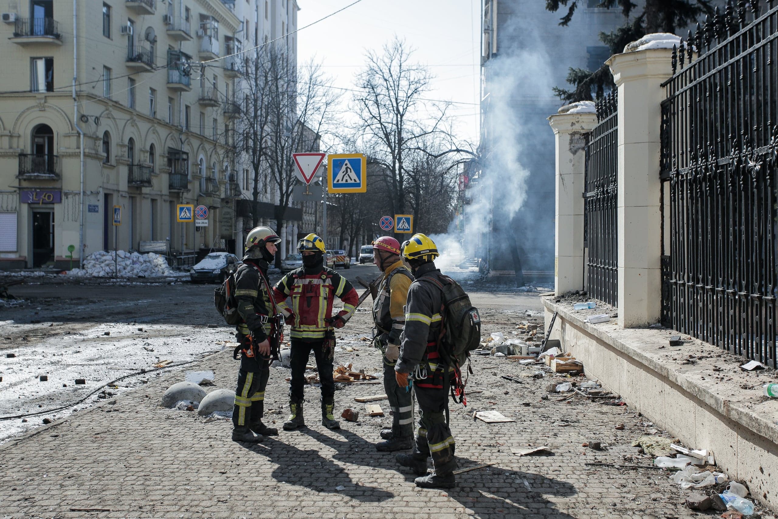 Четыре спасателя стоят на разрушенной городской улице; справа из-за здания поднимается столб дыма.