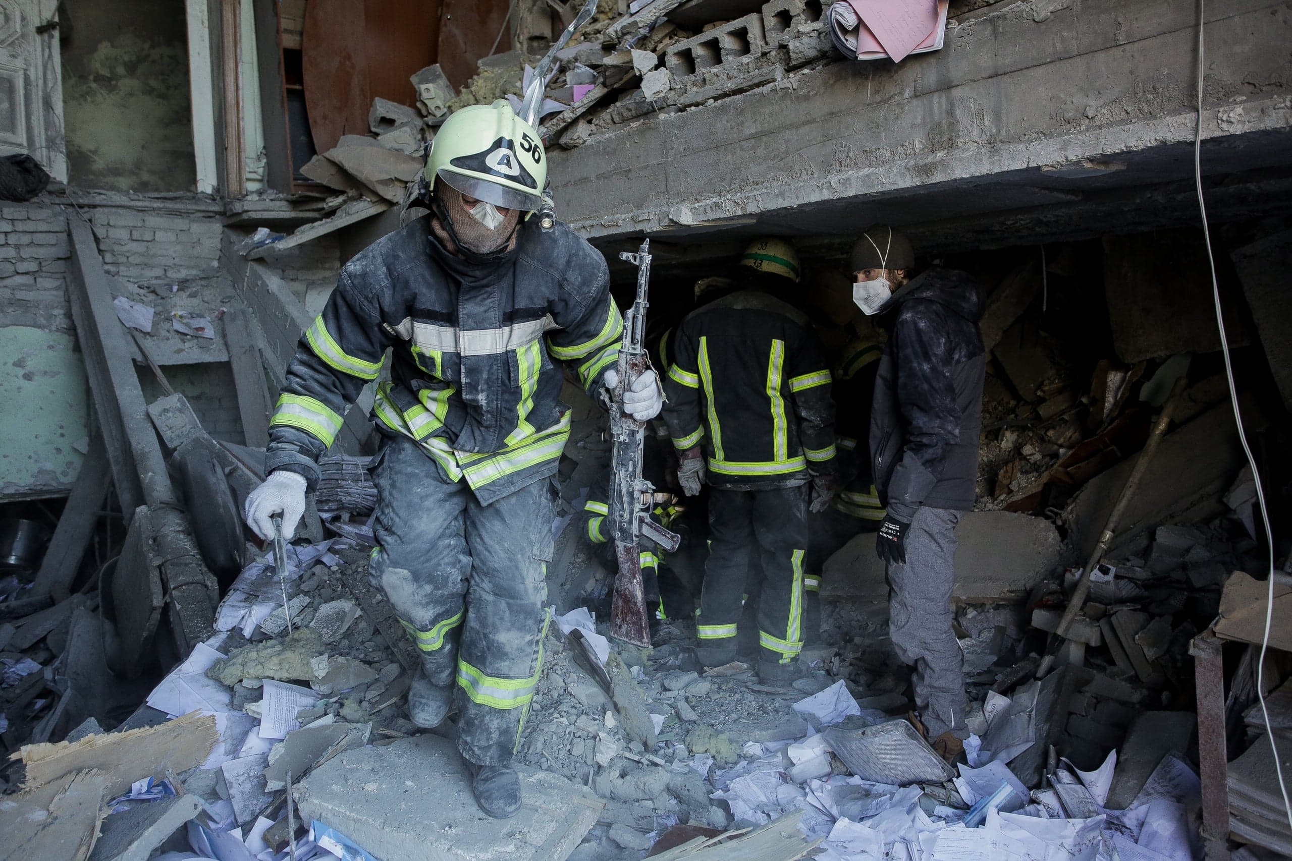 Спасатель в каске с числом "56" и грязной спецодежде держит автомат, стоя среди обломков разрушенного здания; рядом другие спасатели и гражданский в маске.