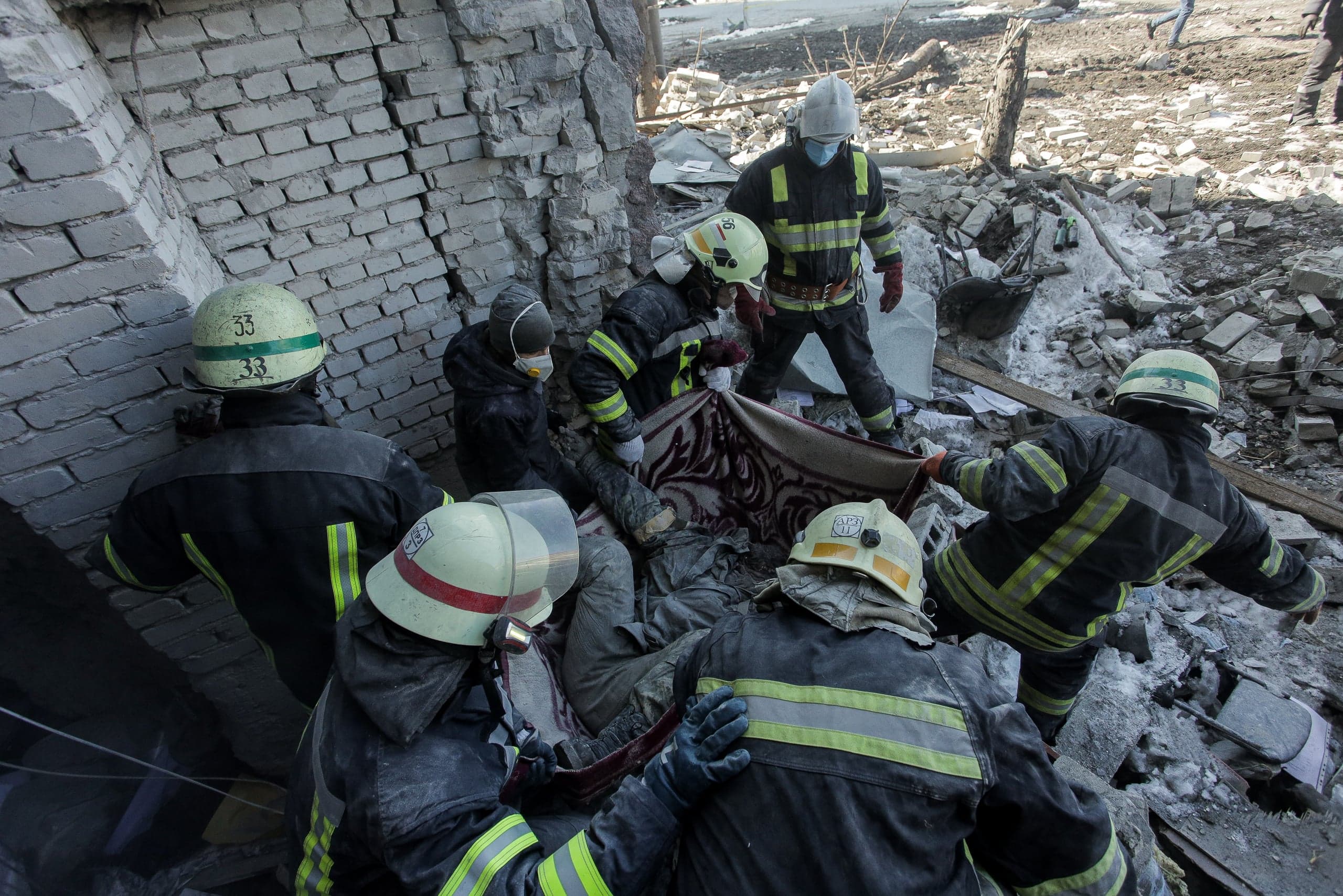 Спасатели извлекают пострадавшего, накрытого ковром, среди руин разрушенного здания.