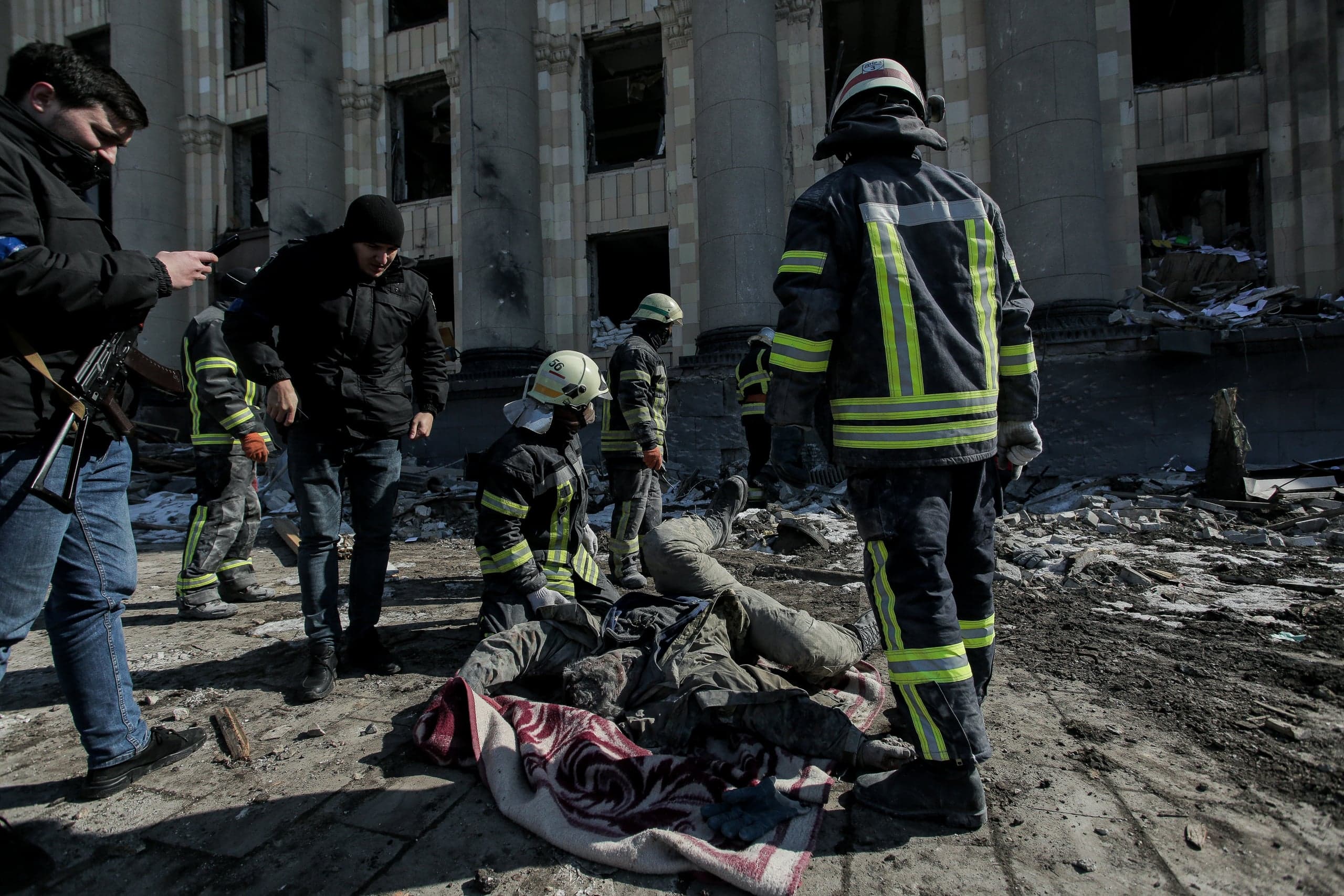 Спасатели и гражданские лица стоят у тела погибшего на фоне разрушенного обстрелом здания.