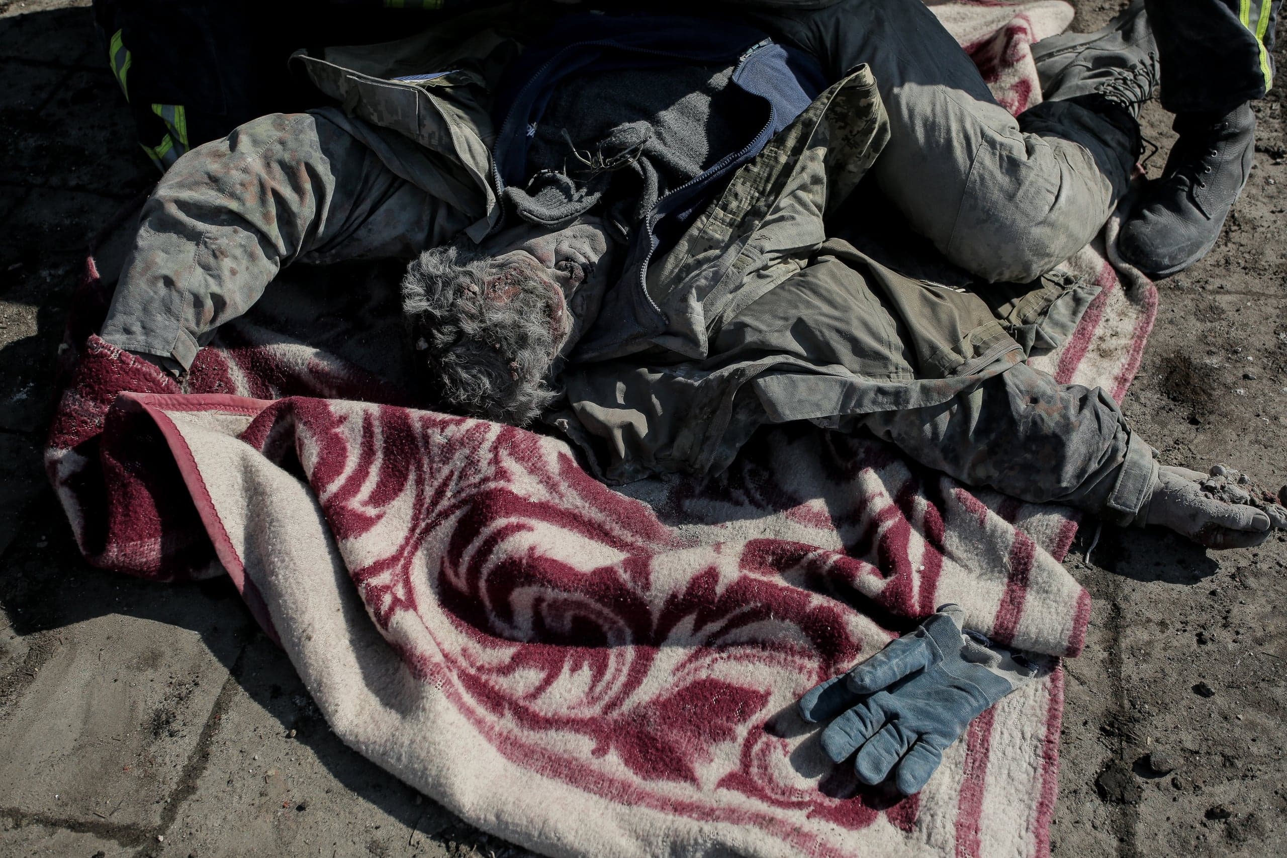 Грязный труп человека лежит на красном покрывале на асфальте, рядом видно ногу стоящего человека 