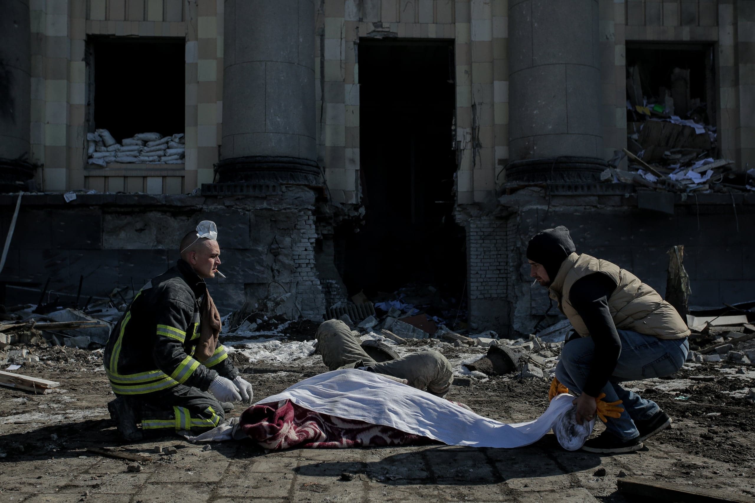 Спасатель и гражданский склонились над телом, накрытым тканью, на фоне сильно разрушенного здания.