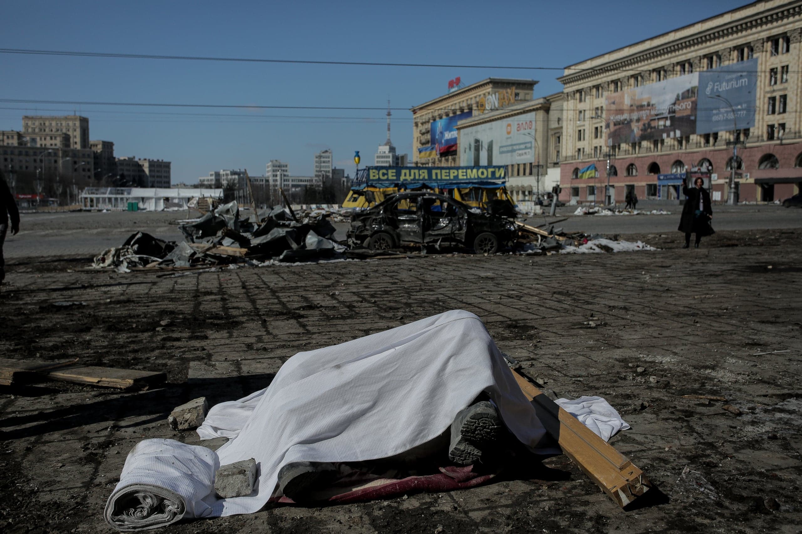 Разрушенная площадь с накрытым белой тканью телом и обломками сгоревшего автомобиля. Вдалеке виден баннер "ВСЕ ДЛЯ ПЕРЕМОГИ".