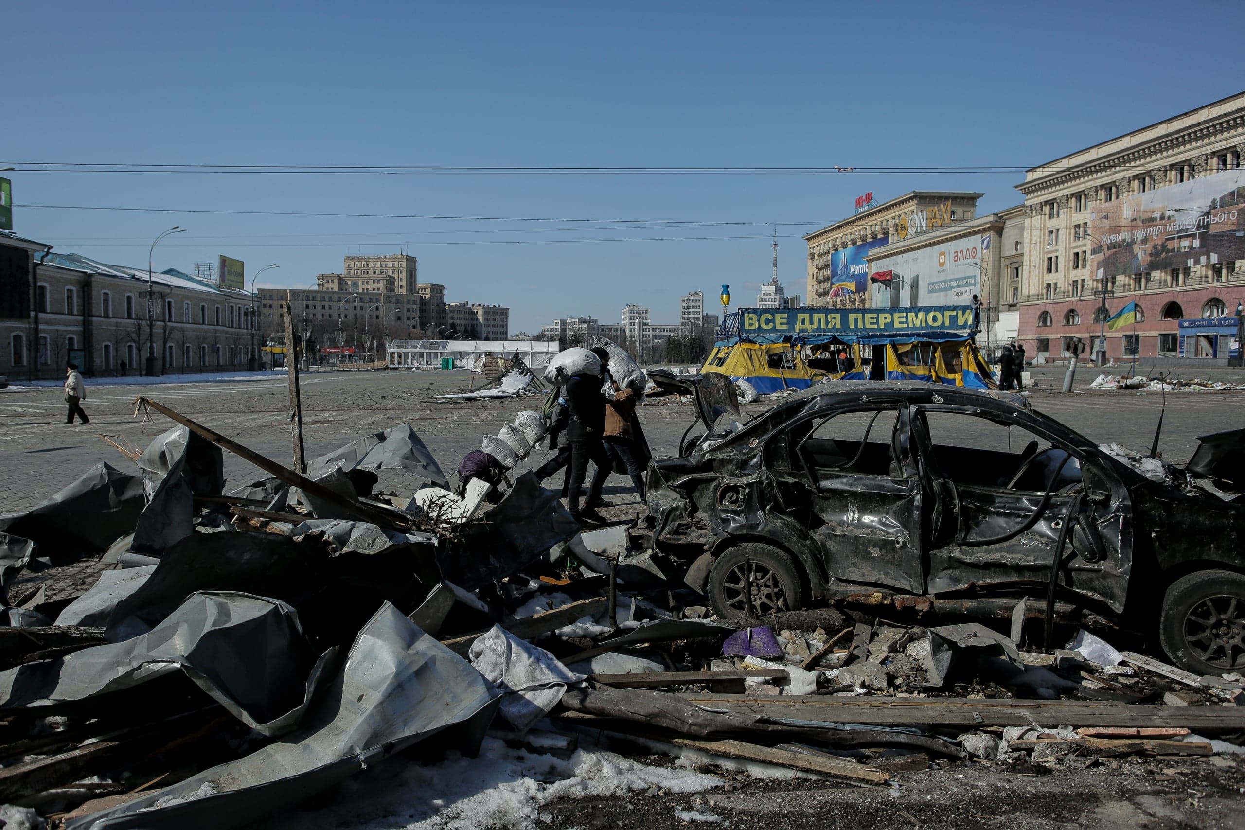 Сгоревшая машина и груды обломков на городской площади. Двое людей несут мешки. На баннере: "ВСЕ ДЛЯ ПЕРЕМОГИ".