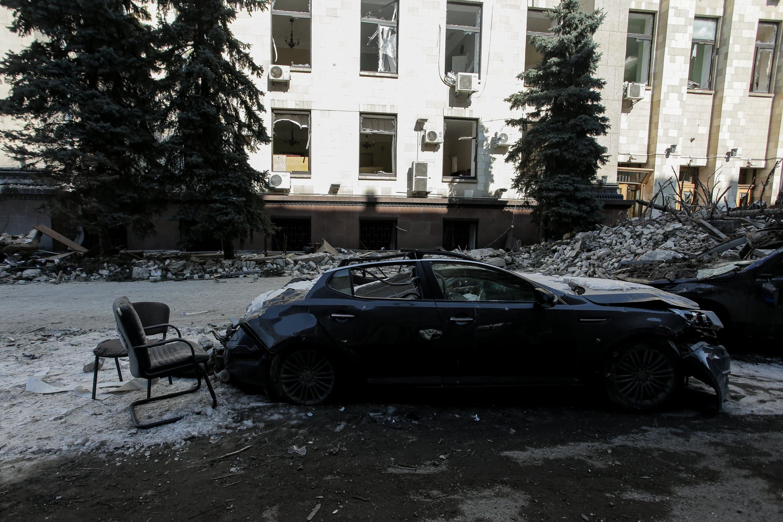 Разрушенный автомобиль среди обломков на фоне поврежденного здания с выбитыми окнами.