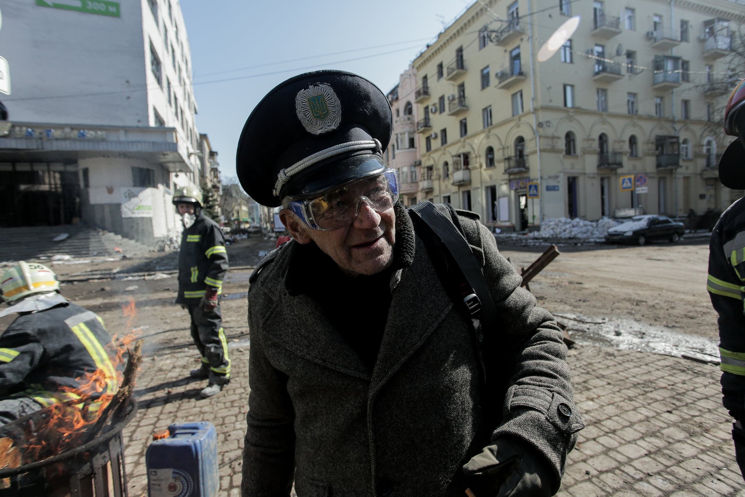 Пожилой мужчина в фуражке, пальто и защитных очках на фоне разрушенной городской улицы с пожарными, один из которых греется у огня в бочке.