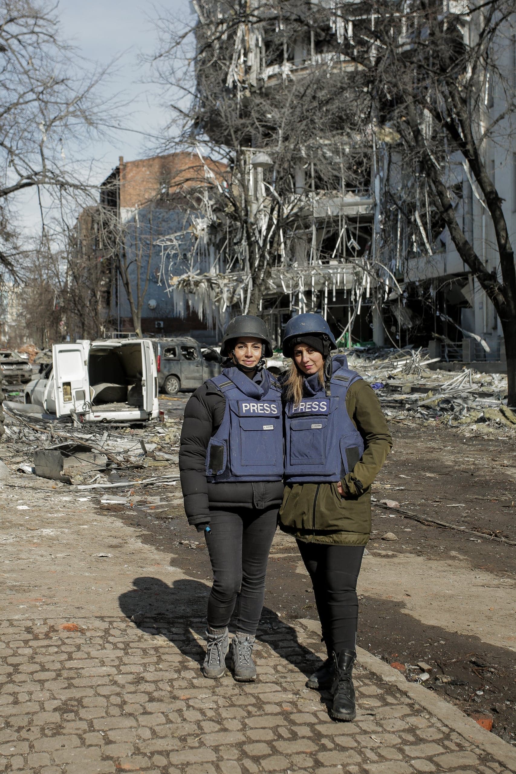 Две журналистки в шлемах и бронежилетах с надписью "PRESS" стоят на фоне разрушенного здания и обломков.