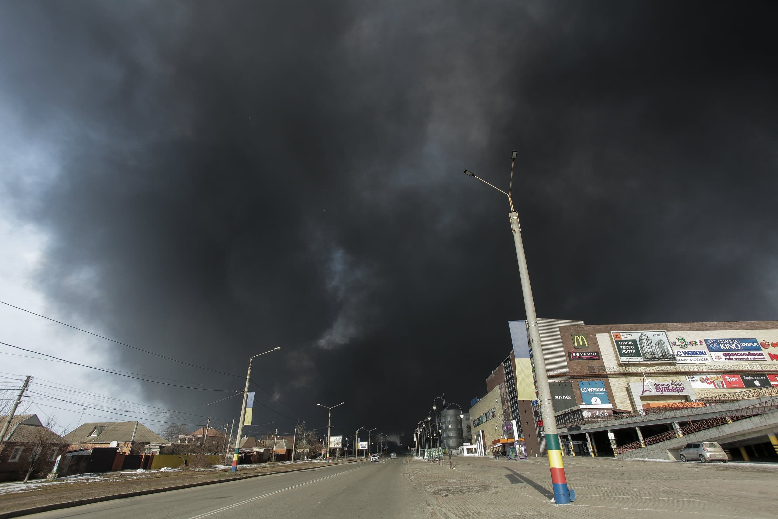 Городская улица, накрытая плотным черным дымом. Справа торговый центр с вывесками, включая логотип McDonald's и надписи "KINO".