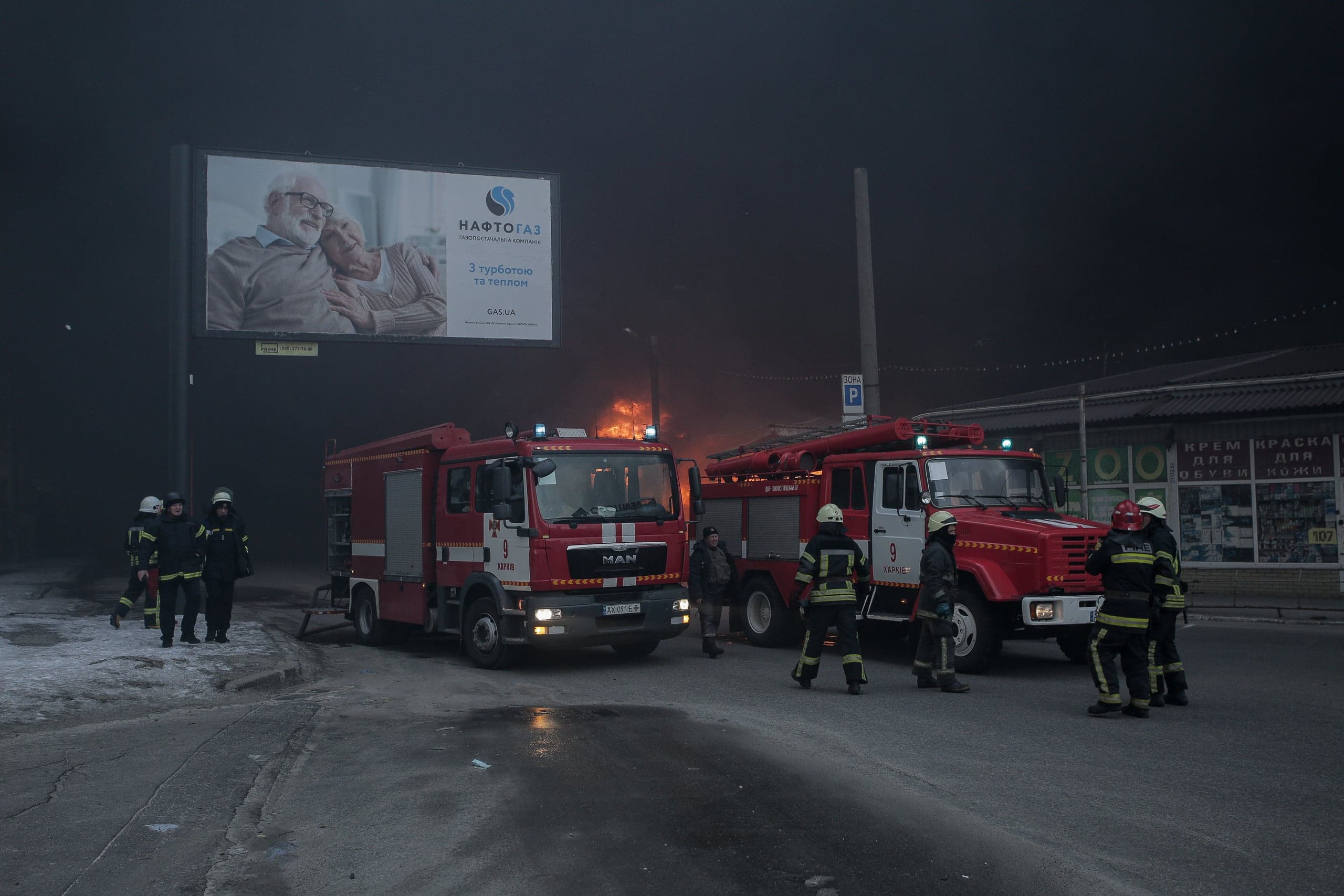 Две пожарные машины и спасатели на ночной улице. На заднем плане виден очаг возгорания. Над сценой рекламный щит с текстом "НАФТОГАЗ".