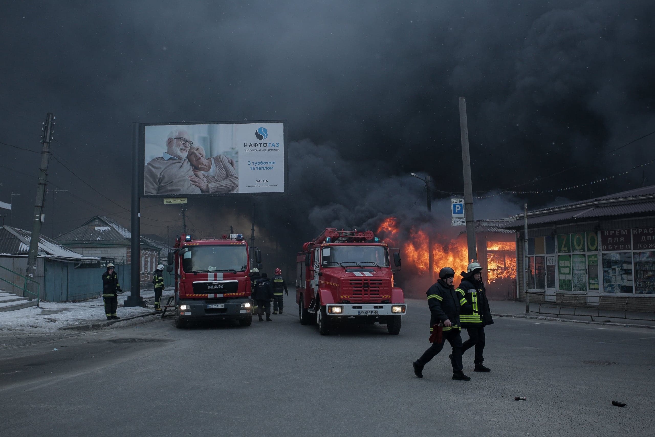 Горящее здание, выпускающее густой черный дым, на городской улице. Два пожарных автомобиля и несколько пожарных на переднем плане. Рекламный щит с текстом "НАФТОГАЗ" и "З турботою про тепло".