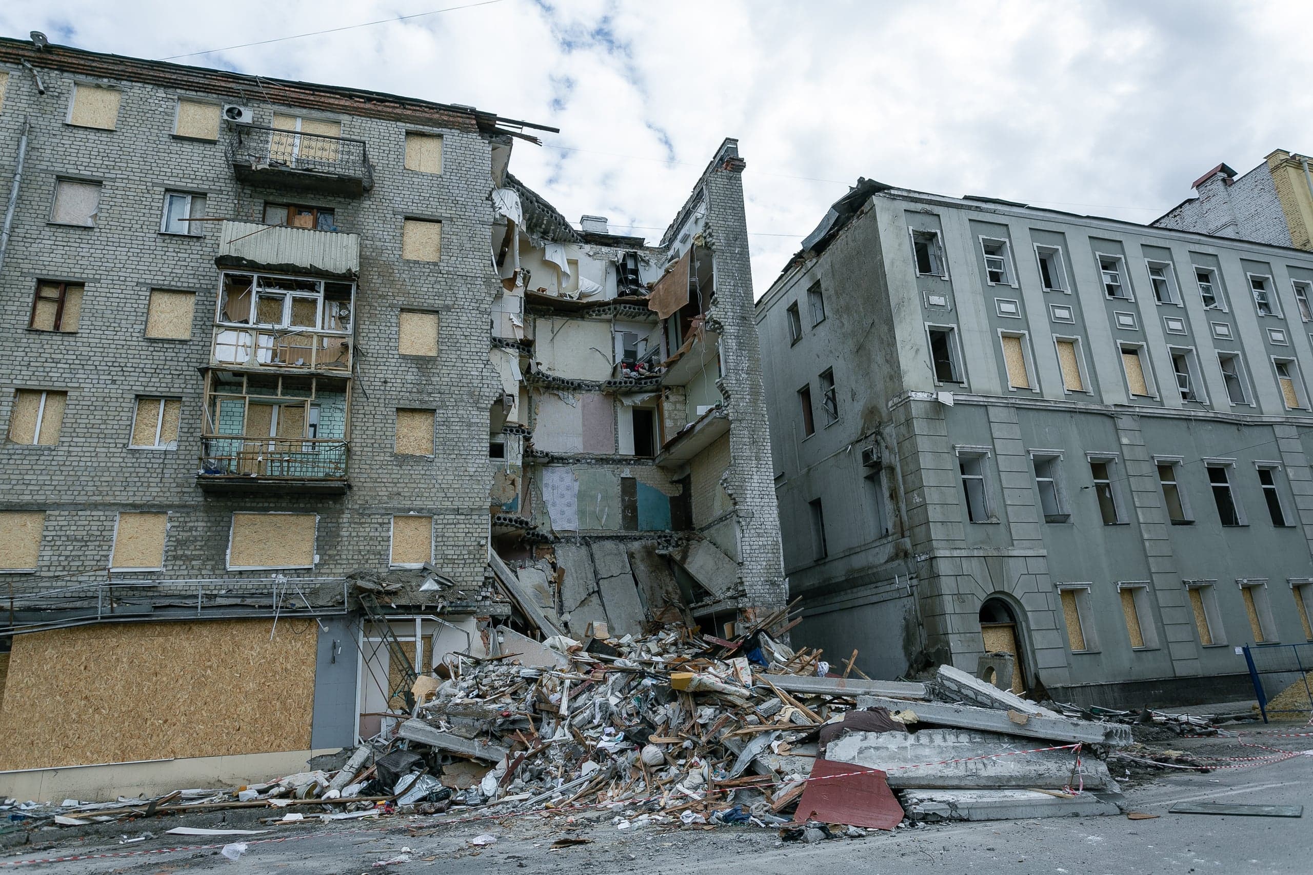 Разрушенный взрывом жилой дом с обрушившейся секцией и грудой обломков на улице. Окна соседних зданий забиты фанерой.