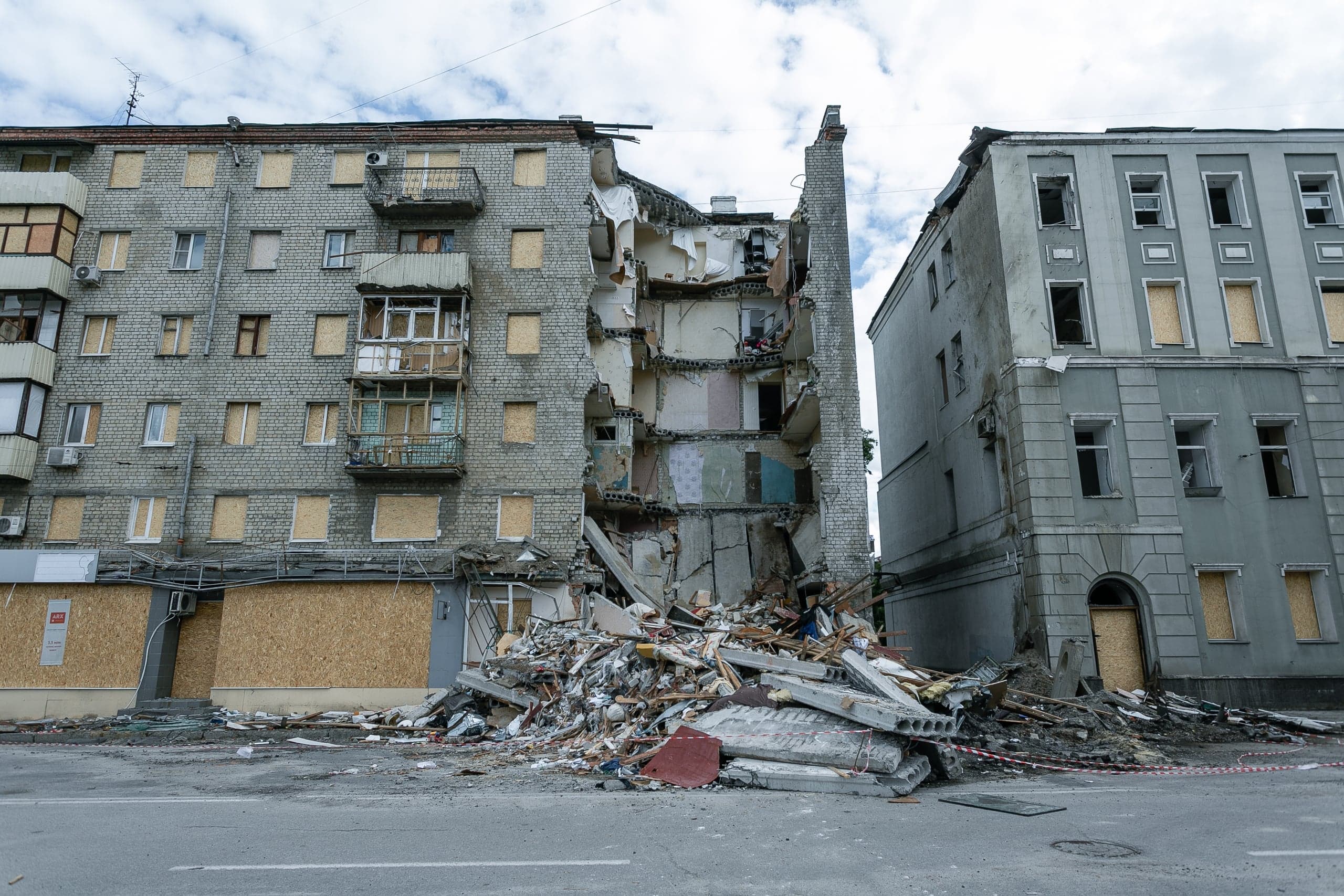 Разрушенный взрывом жилой дом: центральная секция обрушена, обнажая внутренние помещения. Перед зданием лежит большая груда обломков. Окна соседних секций заколочены фанерой.