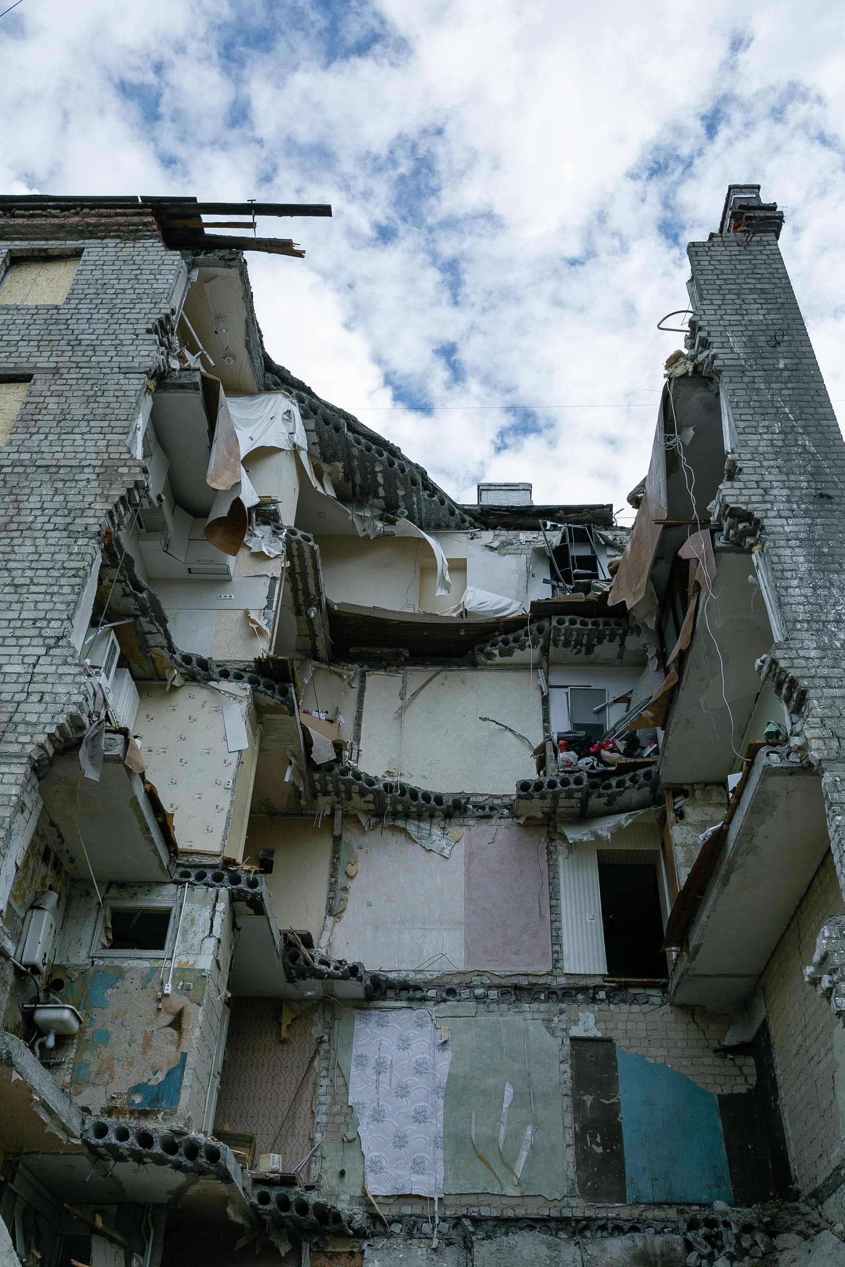 Разрушенный многоэтажный кирпичный дом: центральная часть фасада обрушена, видны поврежденные межэтажные перекрытия и интерьеры квартир.