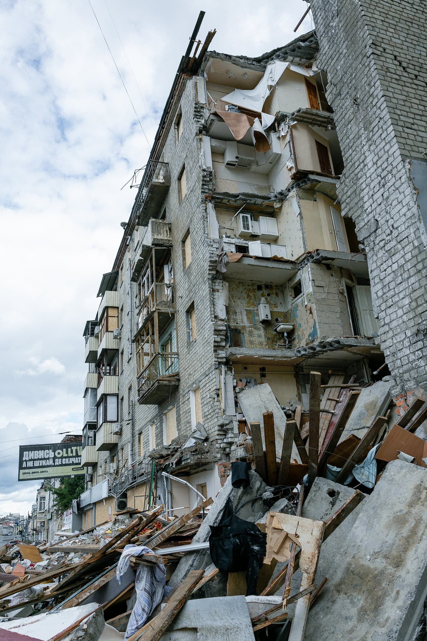 Разрушенный взрывом многоквартирный дом с обрушенным углом и грудой обломков. На заднем плане баннер с текстом "ДИВИСЬ ОЛІТ".