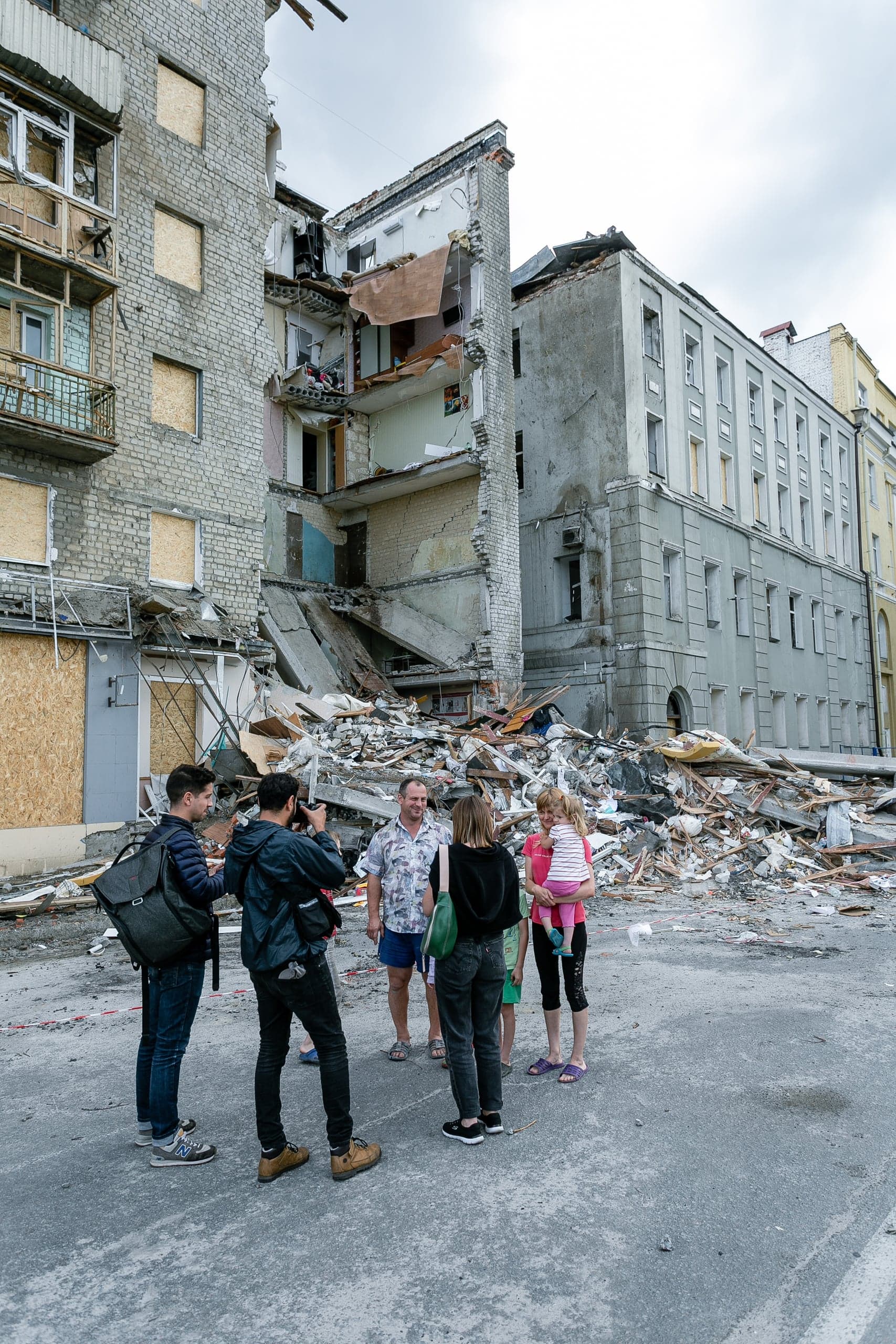 Частично обрушенный многоквартирный дом и груда обломков на улице. Группа людей, включая журналистов и местных жителей с ребенком, стоит перед завалами.