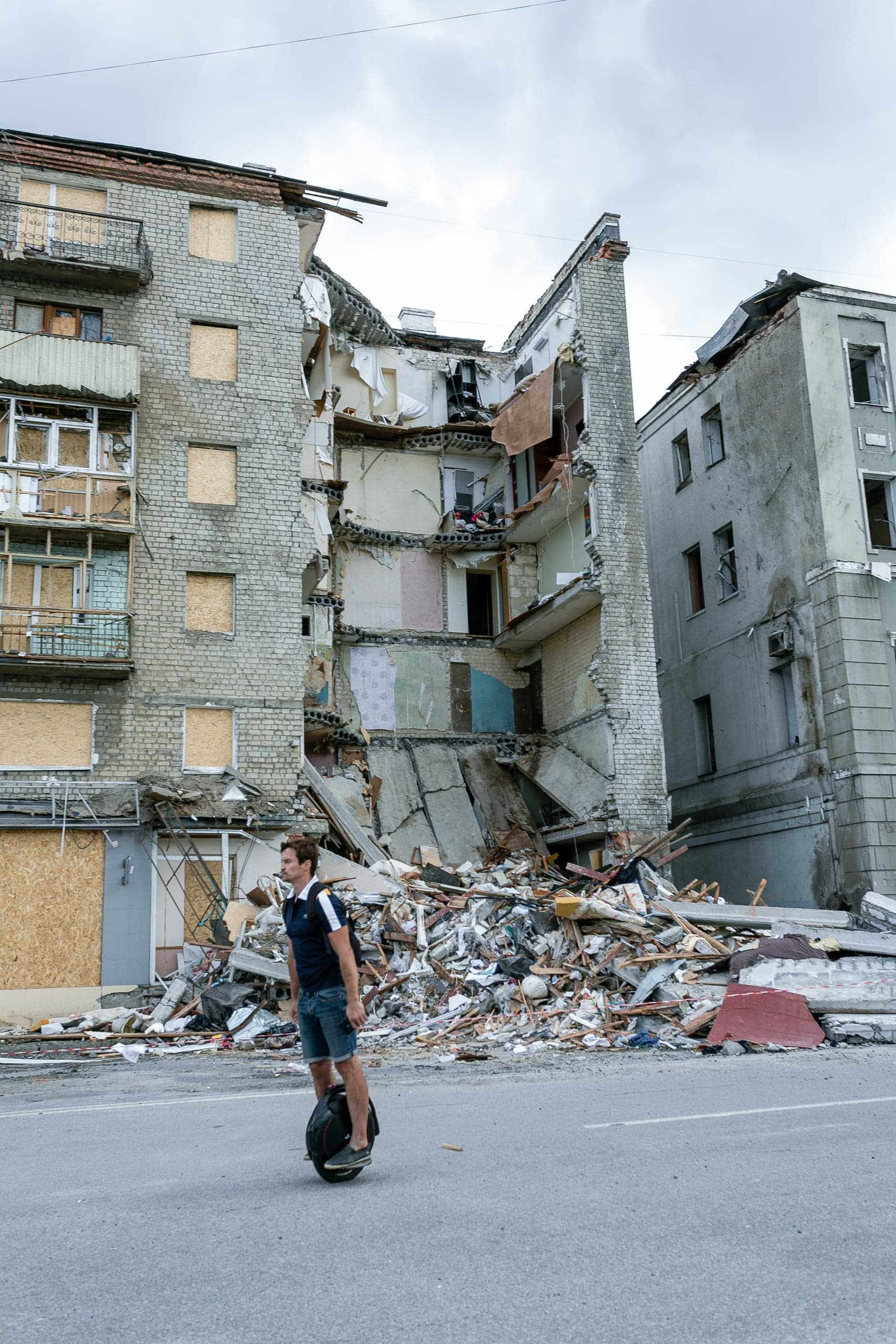 Мужчина на моноколесе едет по улице на фоне частично разрушенного взрывом многоквартирного дома и большой груды обломков.
